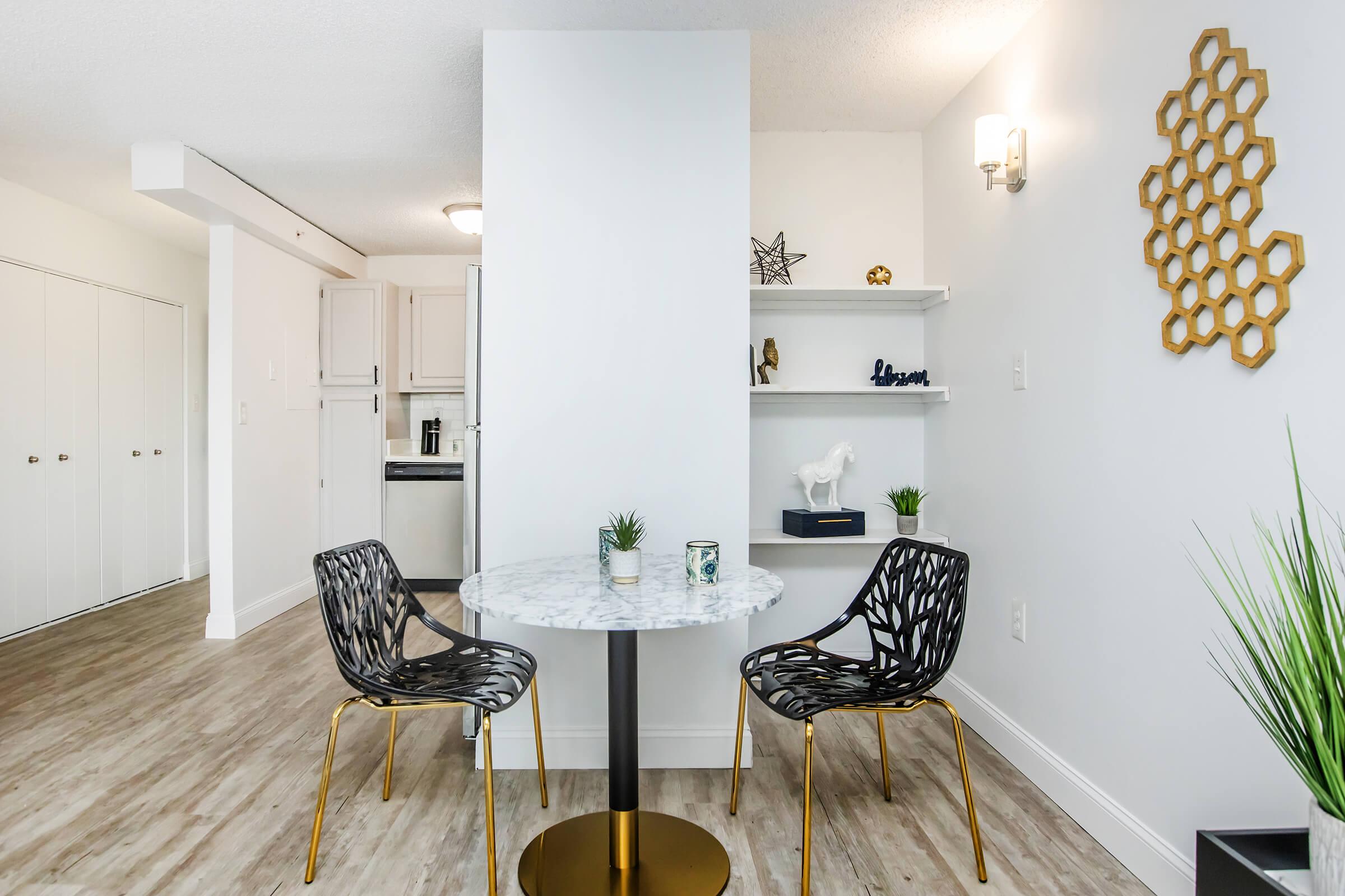 A modern dining area featuring a round marble table with gold accents and two stylish black chairs. In the background, there are light-colored cabinets and a glimpse of a kitchen. A decorative shelf displays small plants and decorative items, while a geometric wall art piece adds a touch of elegance.