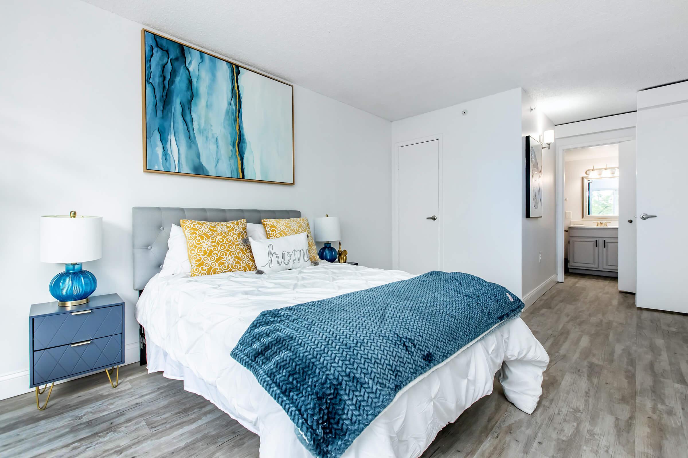 A cozy bedroom featuring a gray upholstered bed with yellow and white decorative pillows, a blue blanket, and a stylish nightstand with a blue lamp. The walls are painted white, and there's a large piece of abstract art above the bed. A bright bathroom area can be seen through an open door.