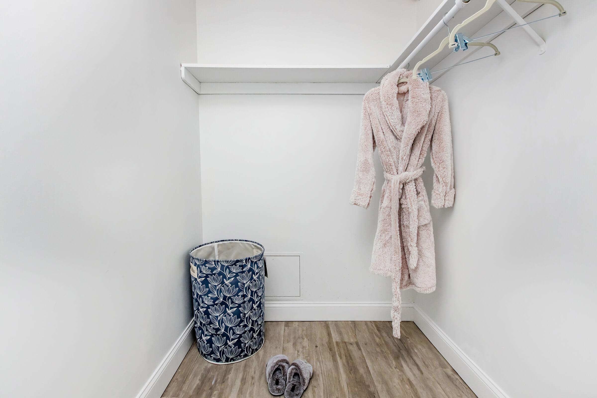A minimalistic closet featuring a pink bathrobe hanging on a white hanger, a woven laundry basket with a floral pattern, and a pair of gray slippers on the wooden floor. The walls are painted white, creating a clean and tidy appearance.