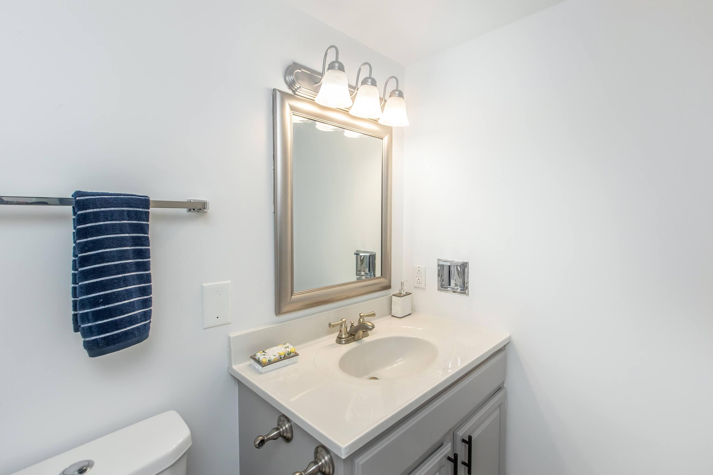 A modern bathroom with a white wall, a sleek white sink with a silver faucet, and a rectangular mirror framed in silver. Above the mirror, there are three light fixtures. A blue striped towel hangs on a towel bar to the left, and a small decorative item is placed on the countertop.