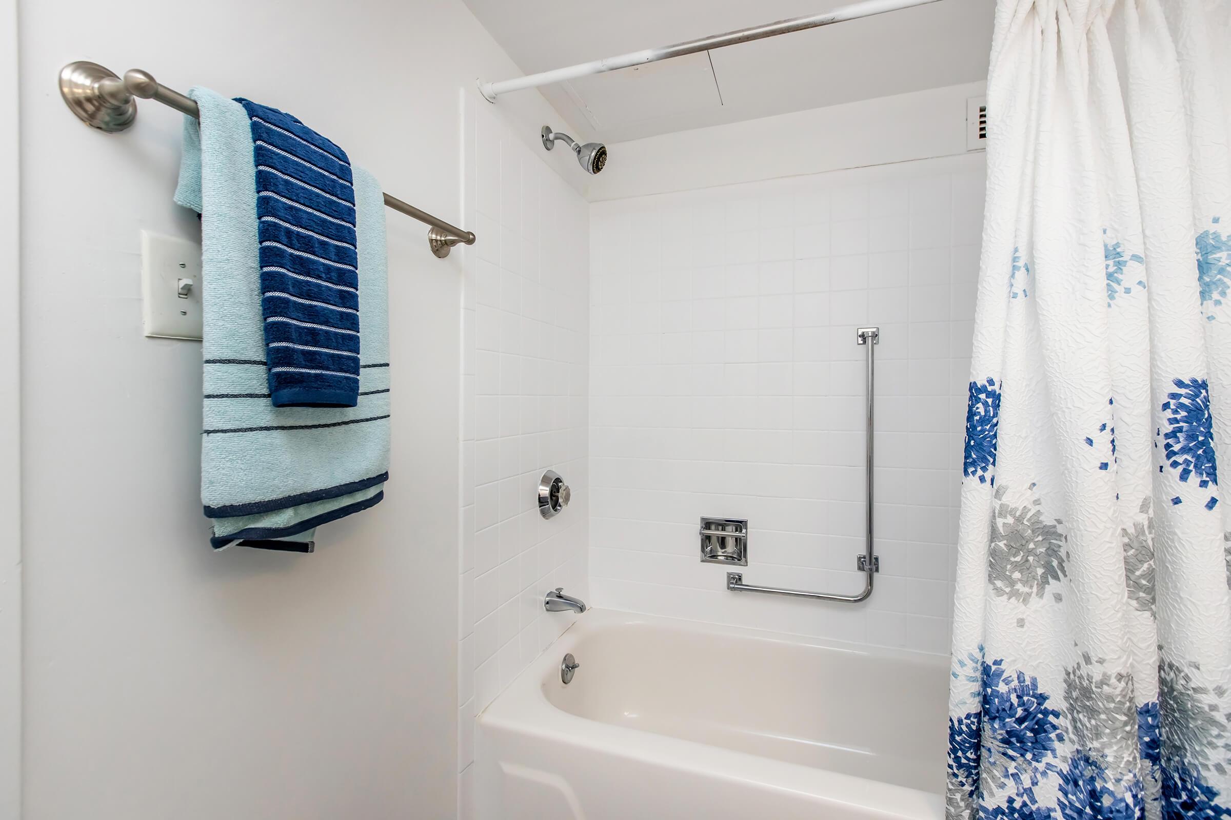A clean, bright bathroom with a white tiled shower area. There is a light blue towel hanging on a chrome towel rack, next to a shower curtain featuring blue and gray floral patterns. A bathtub is visible, with a grab bar and a showerhead above. The overall ambiance is fresh and modern.