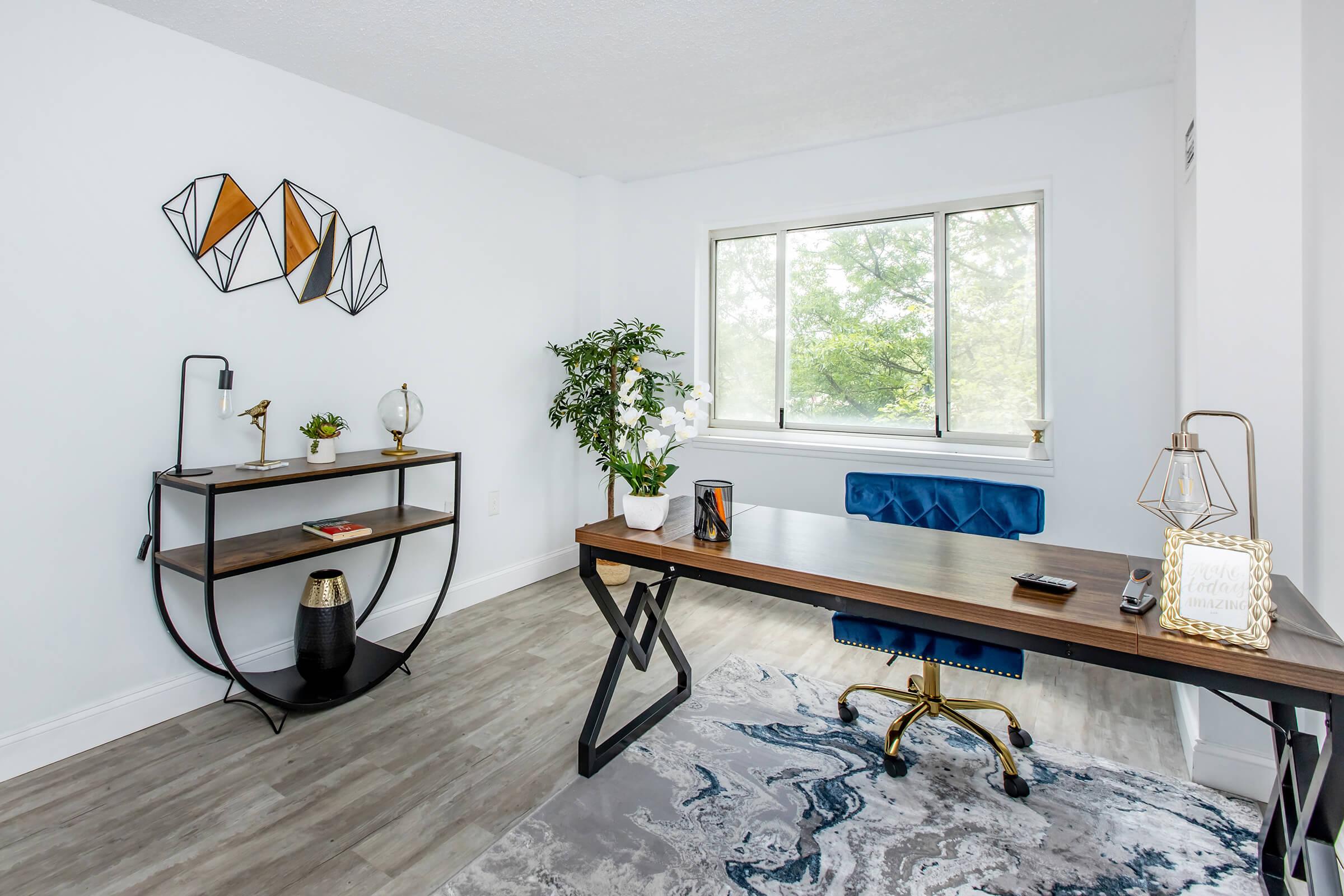 A modern home office featuring a stylish wooden desk with a gold chair and decorative items, a black and gold shelving unit with plants, and a large window allowing natural light. The walls are painted white, and geometric wall art adds a contemporary touch. A rug with blue and gray patterns is underfoot.