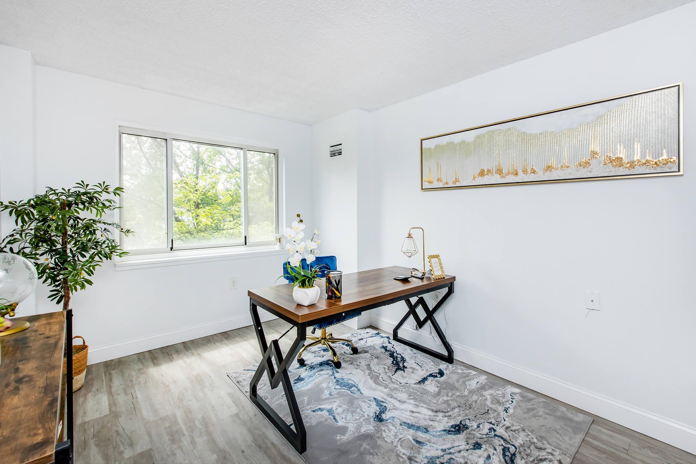 A bright, minimalist home office featuring a wooden desk with a blue chair, a decorative lamp, and a small potted plant. A large window allows natural light to enter, showcasing greenery outside. The floor has light-colored laminated planks, and a large abstract rug adds color to the space. A decorative wall piece hangs above the desk.
