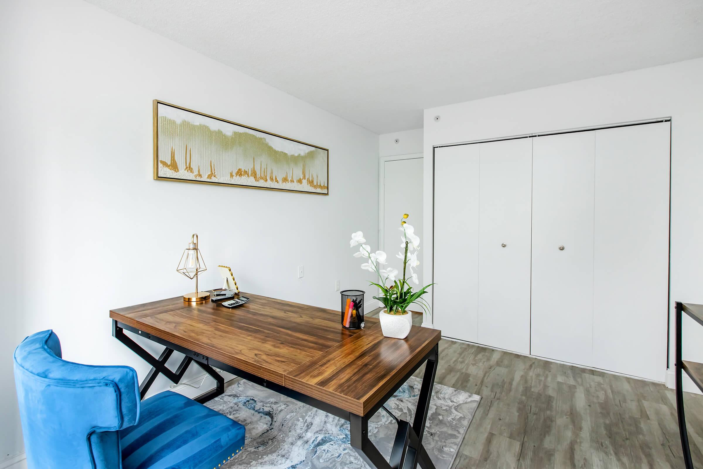 A modern workspace featuring a wooden desk with a lamp, a decorative vase, and a chair. The room has white walls and a large closet with double doors. A minimalist artwork is displayed above the desk, and a patterned rug is placed on the floor, creating a bright and tidy atmosphere.