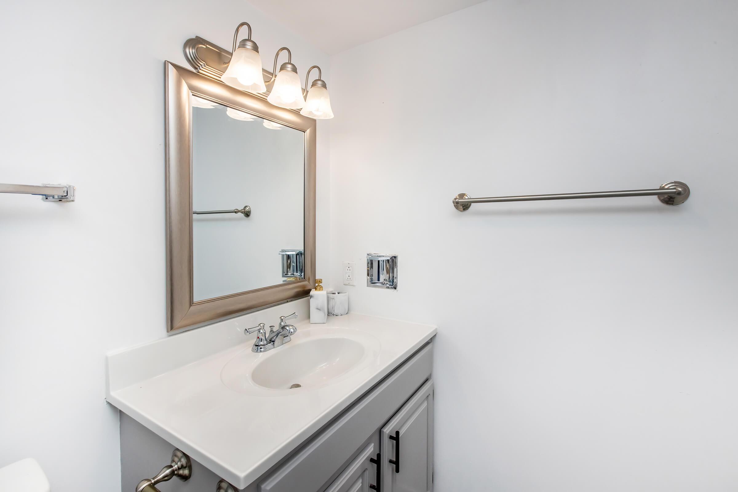 A clean, modern bathroom featuring a white vanity with a sink, a wall-mounted mirror above it, and three light fixtures. The walls are white, and there's a towel rack nearby. The overall ambiance is bright and minimalist.