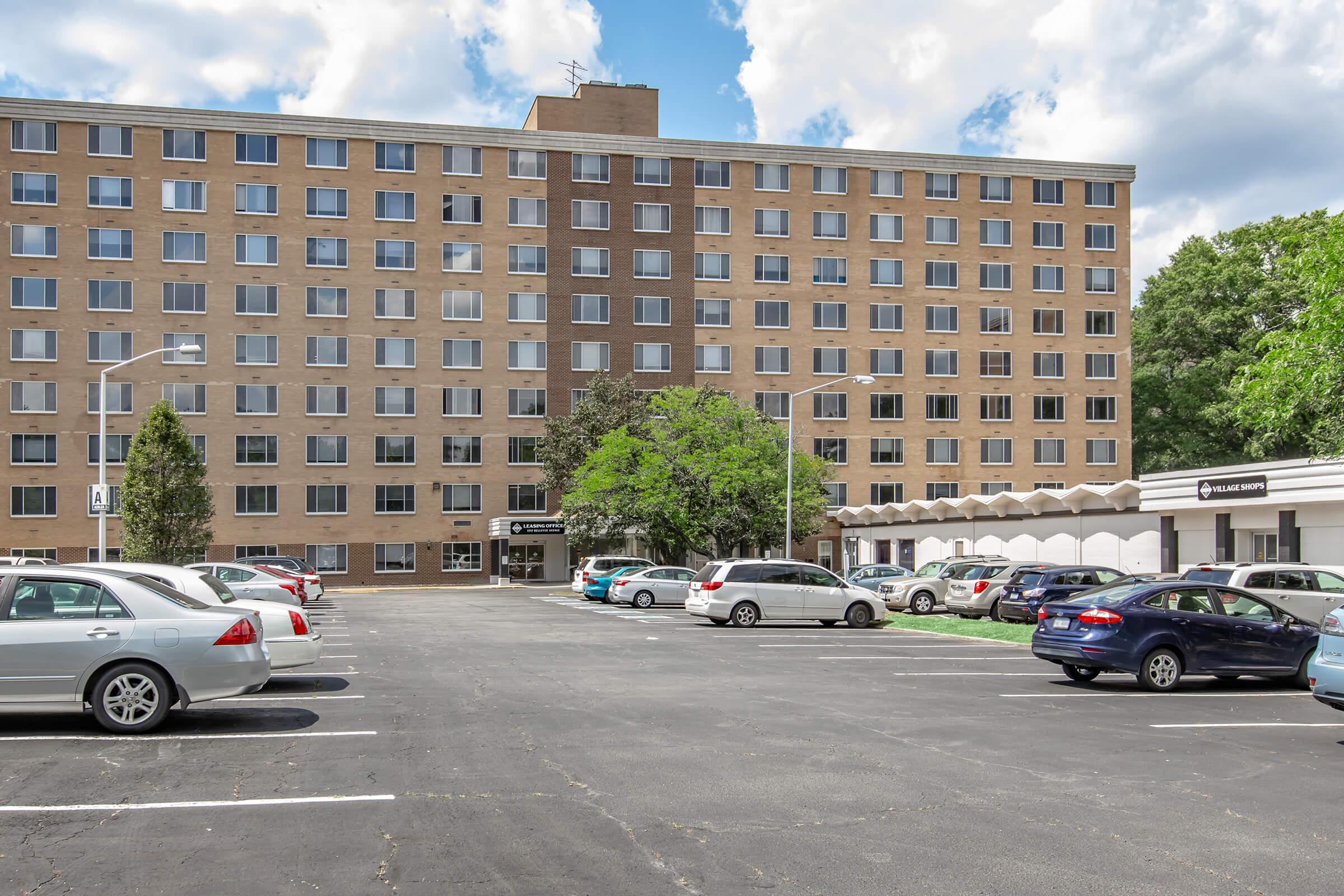 A large, multi-story building with several windows, surrounded by a parking lot filled with cars. In front of the building is a landscaped area with a tree. The sky is partly cloudy, contributing to a bright atmosphere. The scene appears to be a residential or commercial complex.