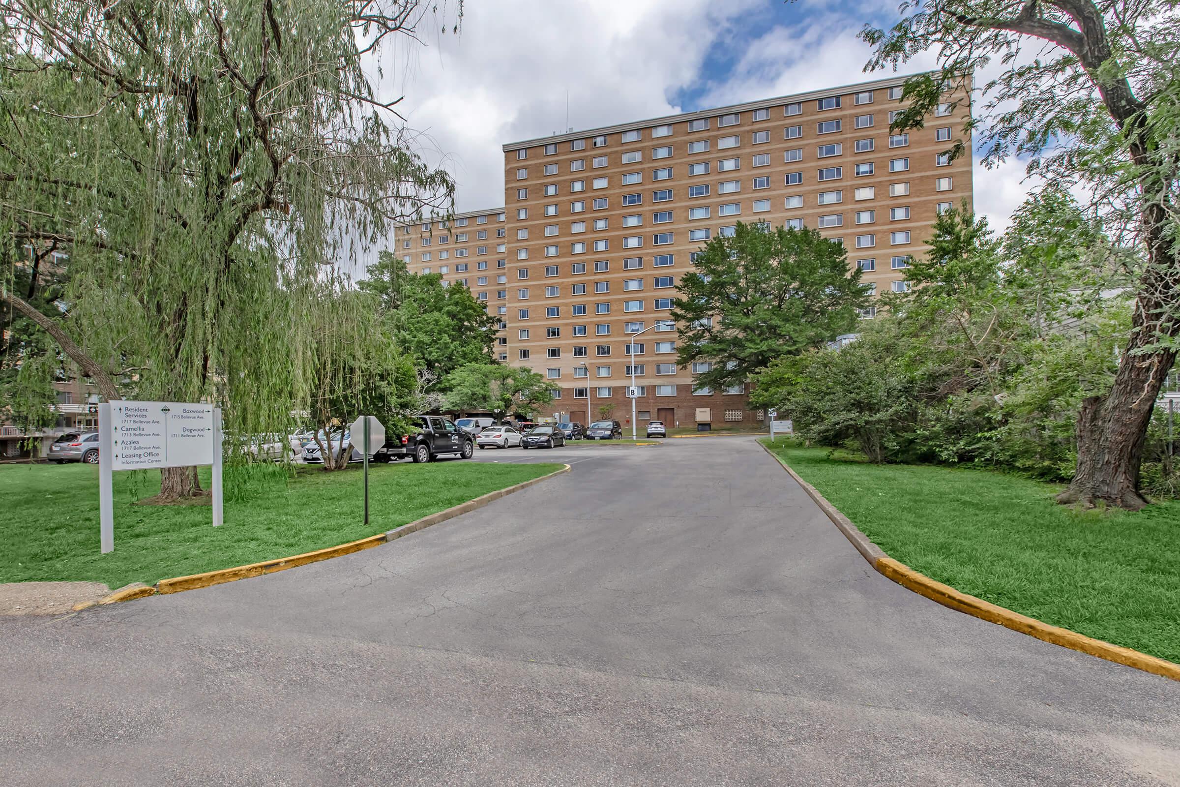 A wide pathway leads to a multi-story brick building surrounded by greenery. The building features numerous windows and is situated near parked cars and trees. A sign is visible on the left, indicating directions or information about the location. The sky is partly cloudy, adding to the serene atmosphere.
