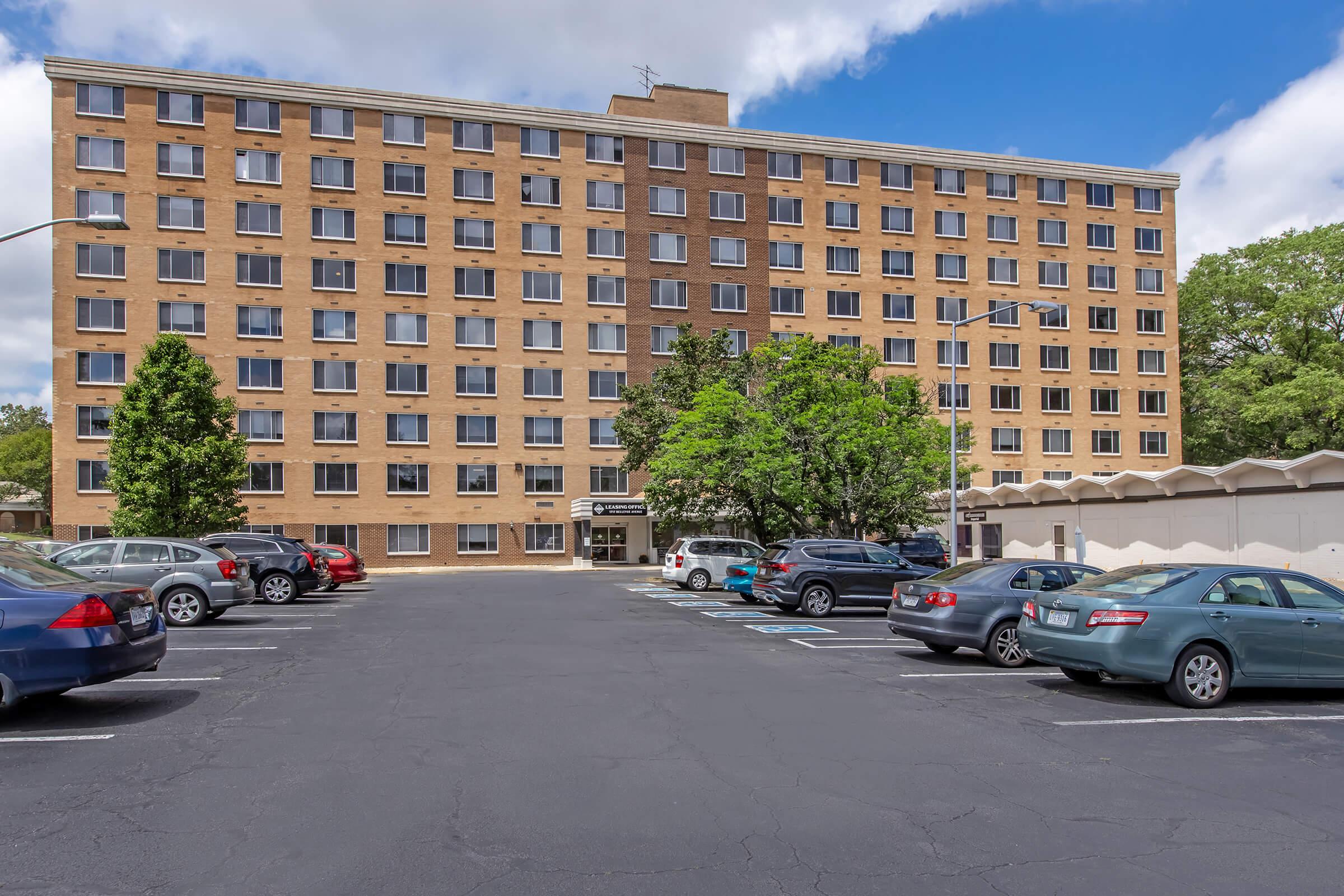 A large, multi-story brick building with multiple windows set against a partly cloudy sky. In the foreground, there is an asphalt parking lot filled with parked cars of various colors. Trees are located near the building entrance, providing some greenery to the scene.