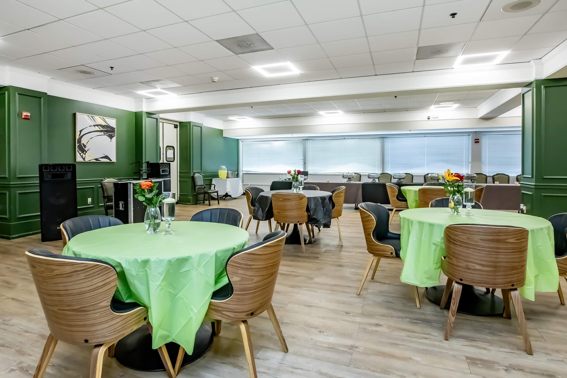 Brightly lit dining area featuring several round tables with green tablecloths. Each table is decorated with small flower vases. The walls are painted dark green, and there are large windows providing natural light. A snack or serving area is visible in the background. The space is inviting and well-organized.