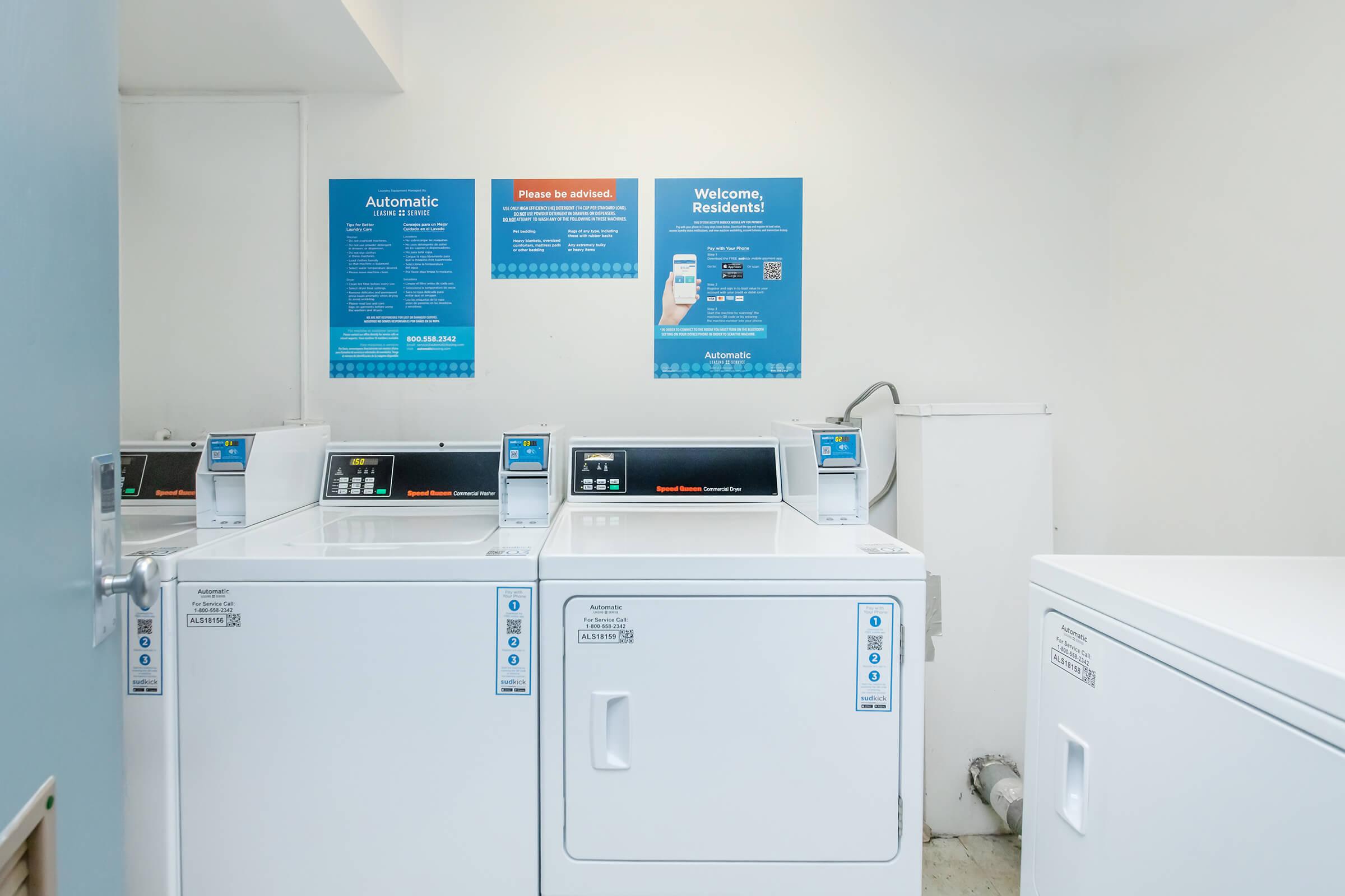 A clean laundry room featuring three white washing machines and a white dryer. Two informational blue posters are mounted on the wall, providing instructions and welcome messages for residents. The space is well-lit and organized, with a plain floor and a door leading to the outside.