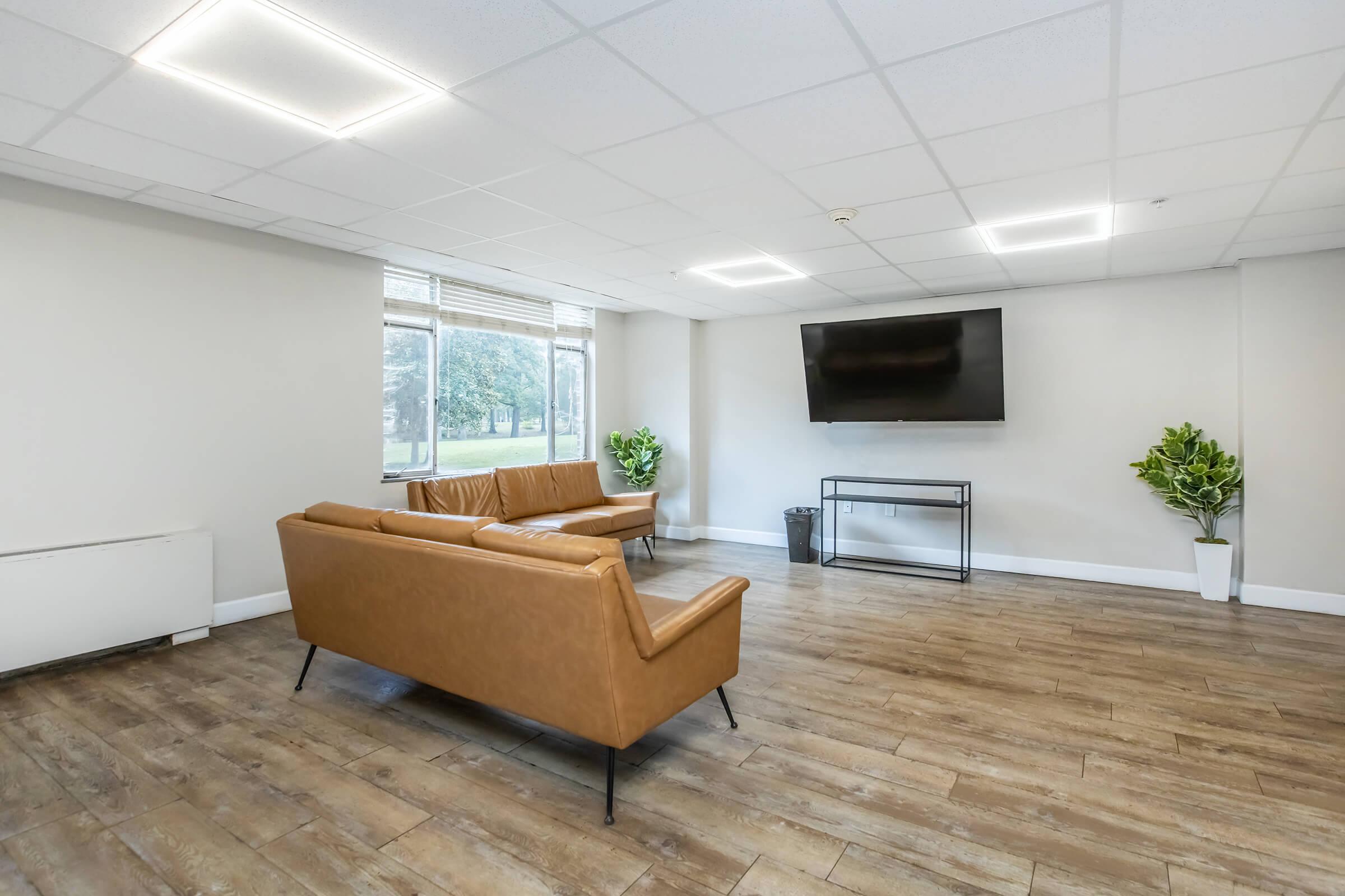A spacious interior room featuring a brown leather sofa, a glass TV stand, and a flat-screen television mounted on the wall. Large windows allow natural light to enter, and there are two potted plants in the corners. The floor is made of wooden planks, contributing to a modern and minimalistic aesthetic.