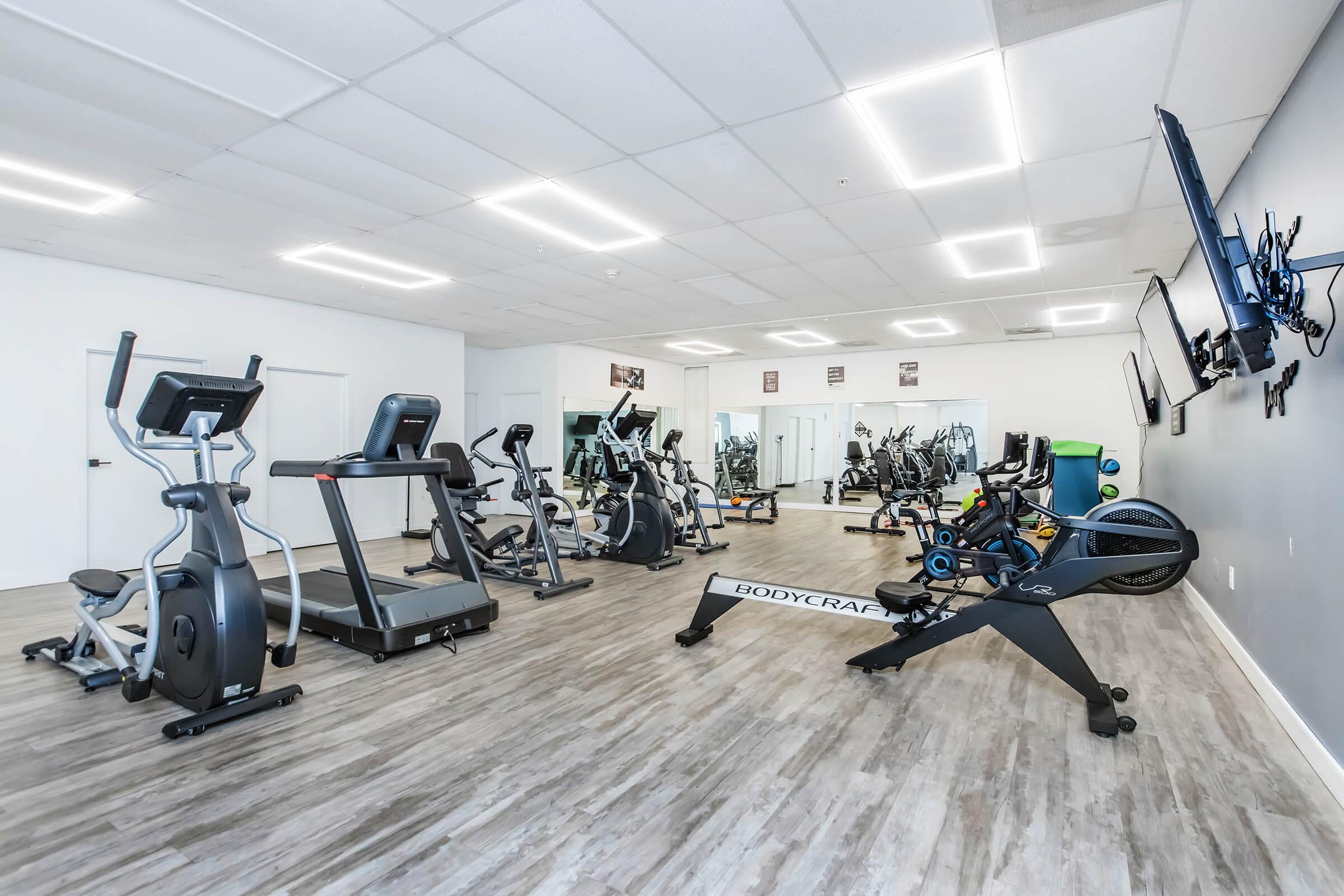 A bright and spacious gym with various exercise equipment including treadmills, ellipticals, and stationary bikes. The room features large windows and a modern design, with multiple screens mounted on the walls displaying workout information. The flooring is a light wood color, contributing to a clean and inviting atmosphere.