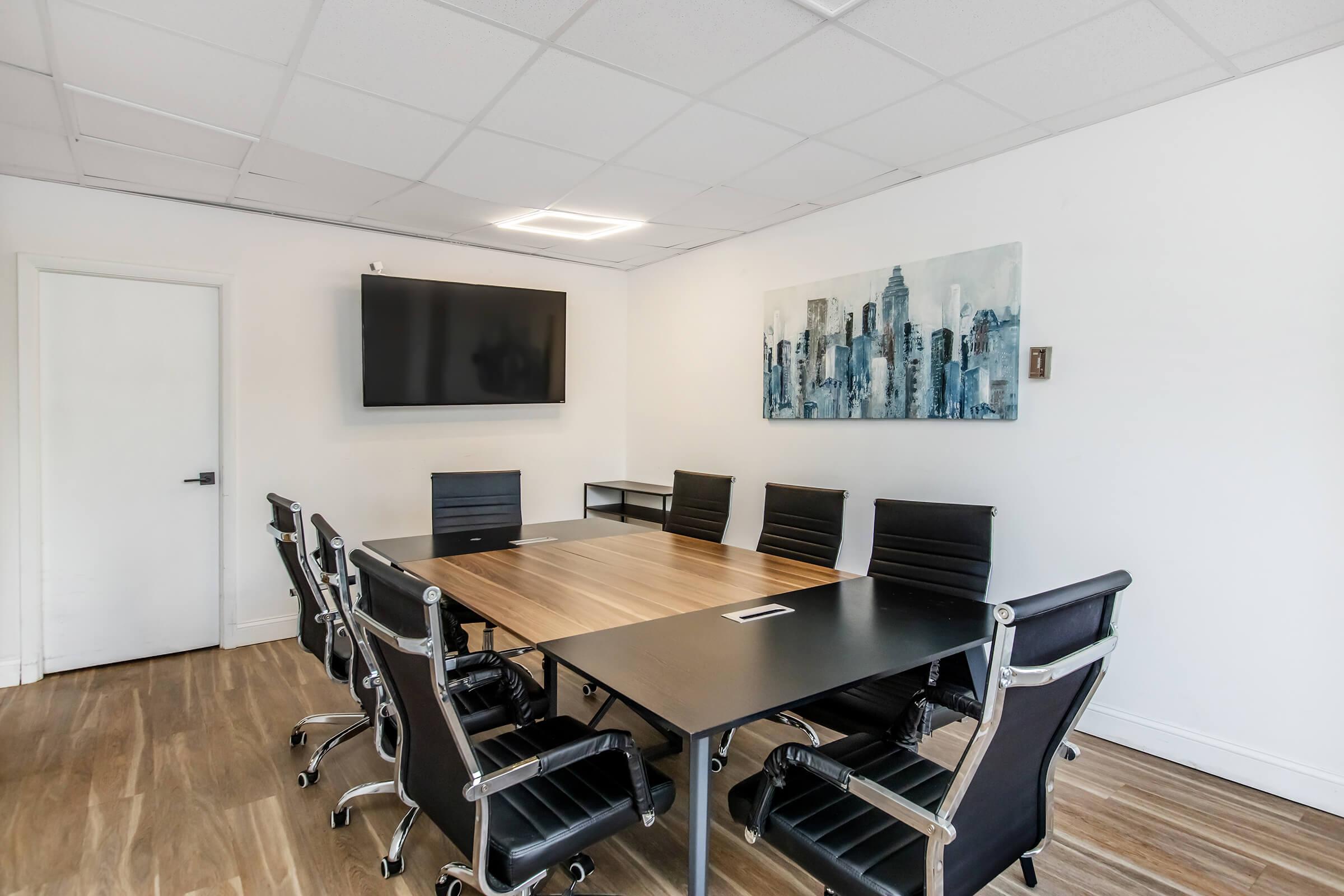 A modern conference room featuring a large wooden table surrounded by black leather chairs. There is a wall-mounted TV on one side and a cityscape artwork on another. The room has bright lighting and a minimalist design, with white walls and wooden flooring. A door is visible in the background.