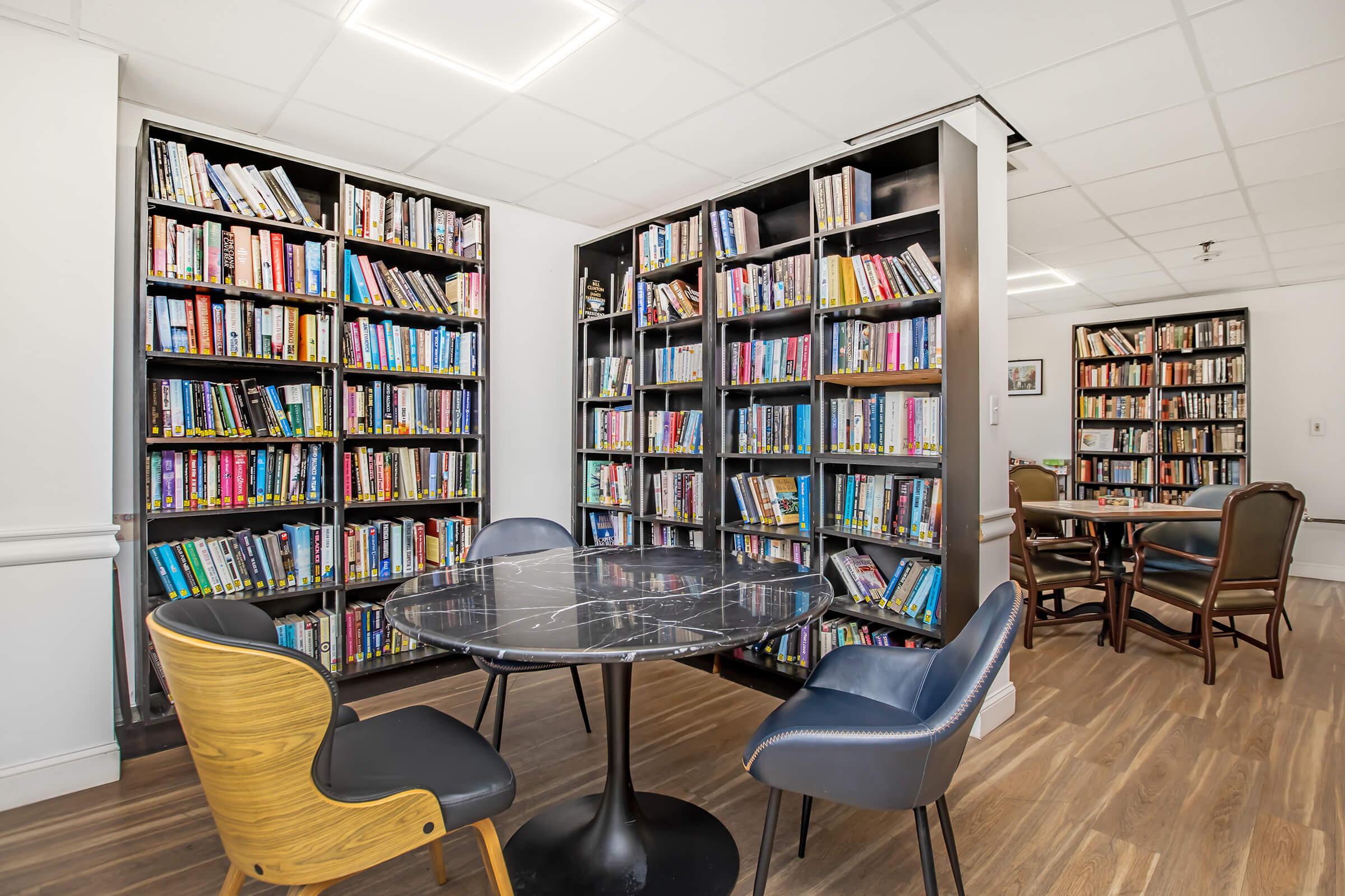 A cozy reading area featuring two bookshelves filled with various books, a round black marble table with four chairs, and a well-lit atmosphere. The space is designed for relaxation and reading, with wooden flooring and a warm ambiance.