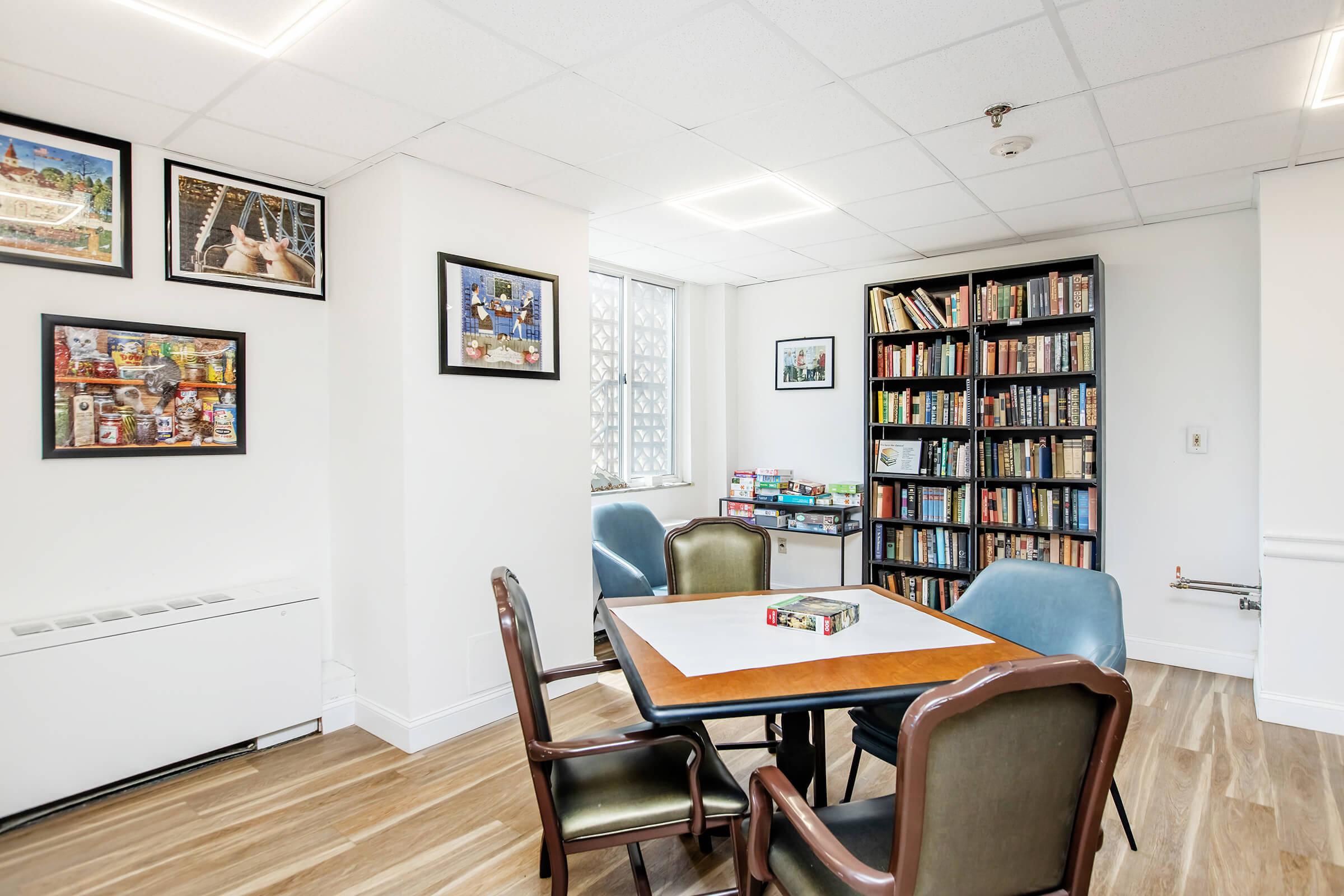 A bright and modern reading or gathering room featuring a table surrounded by four chairs. One wall has a bookshelf filled with books, while another wall showcases framed pictures. The space has large windows allowing natural light and a clean, contemporary design with hardwood flooring.
