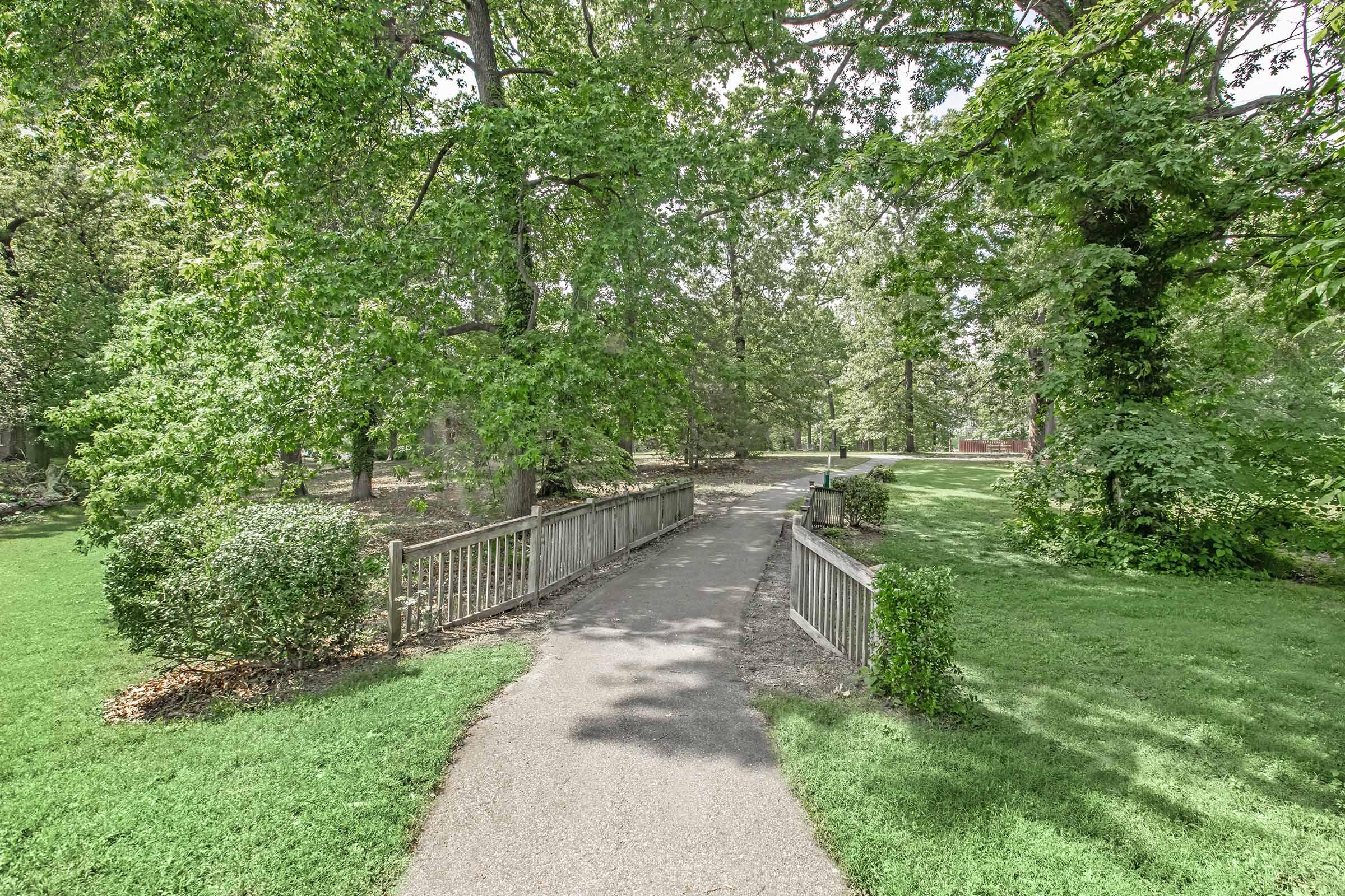 A peaceful park scene featuring a winding pathway surrounded by lush greenery. The path is framed by trees and shrubs, leading to an open area in the distance. Sunlight filters through the leaves, creating a serene and inviting atmosphere perfect for a leisurely walk or relaxation in nature.