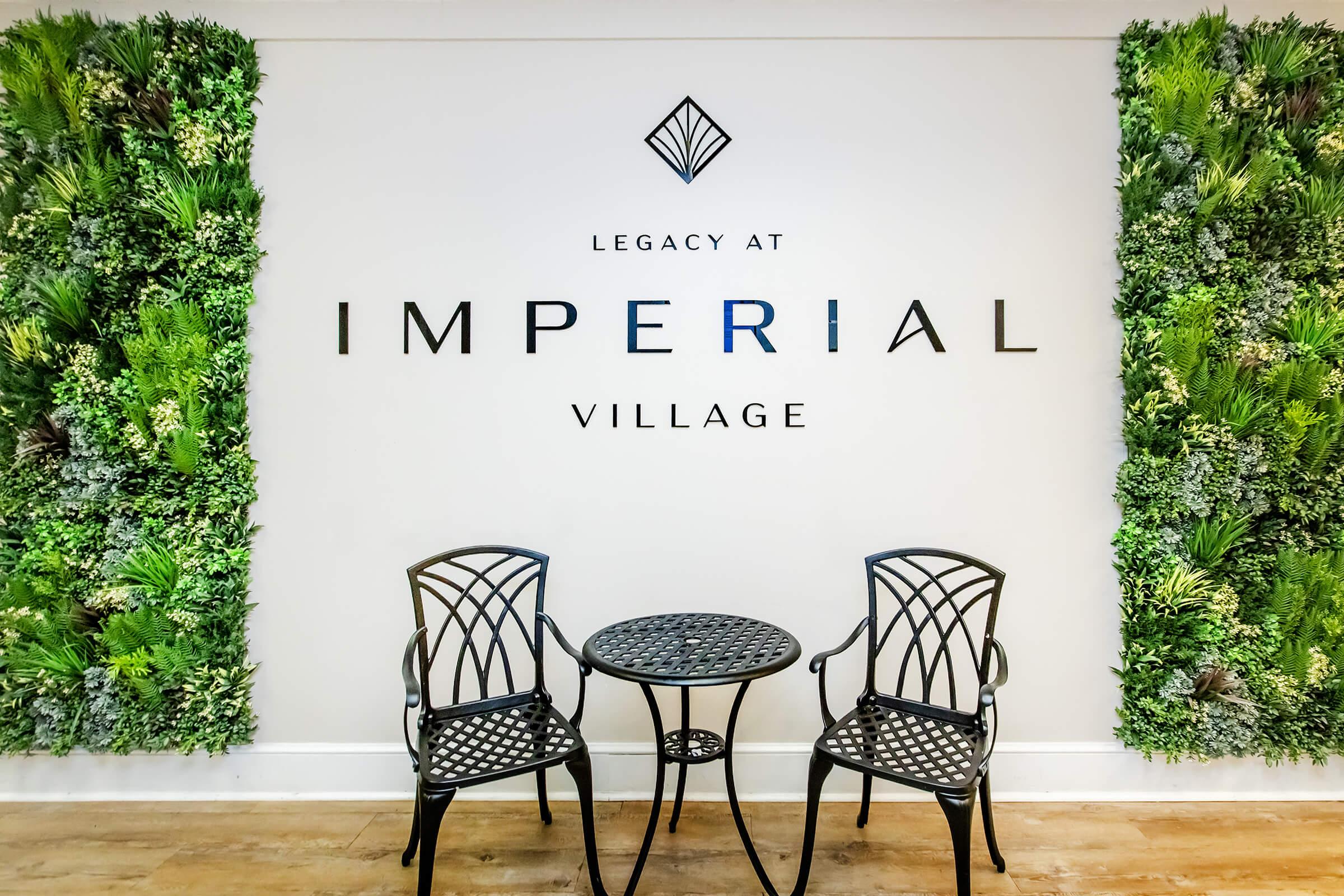 A stylish indoor seating area featuring two black metal chairs and a small round table in front of a wall adorned with vibrant green plants. The wall prominently displays the text "Legacy at Imperial Village" in large, elegant letters.