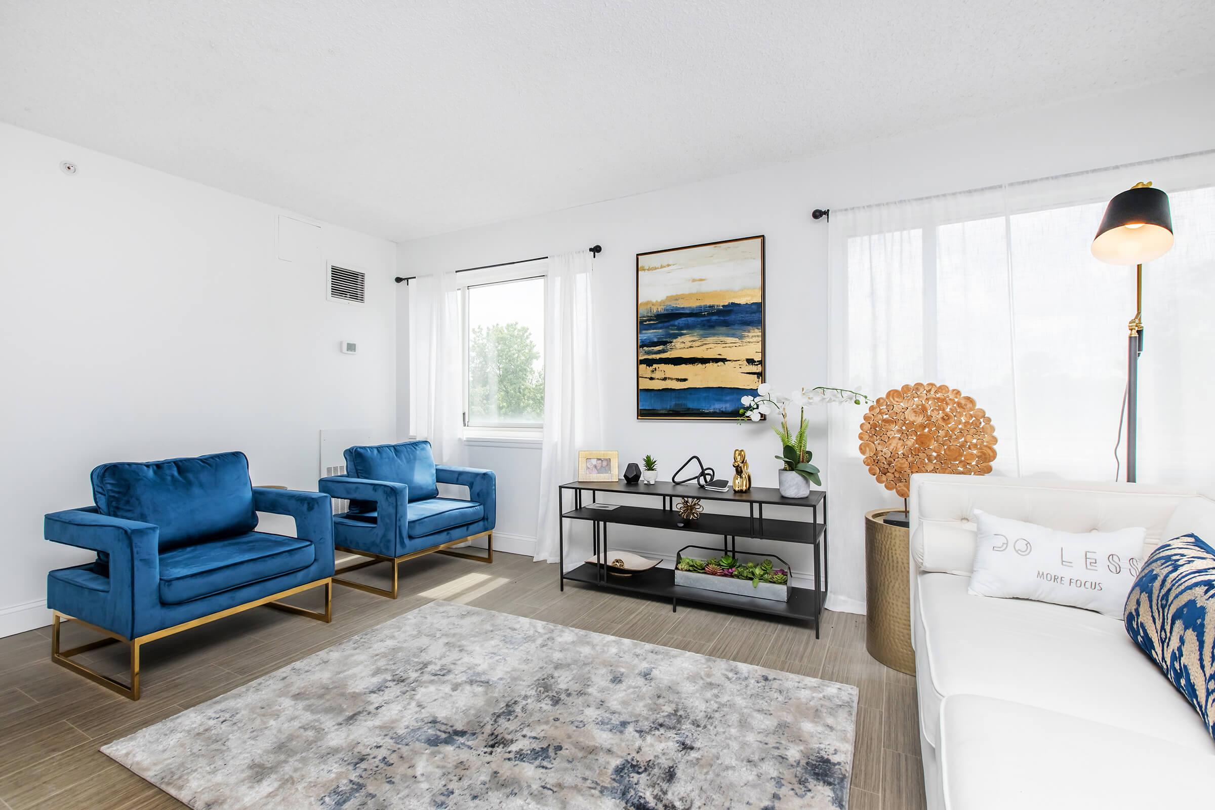 A bright, modern living room featuring two blue velvet armchairs, a white couch, a decorative coffee table with plants and decor, and a large rug. Large windows allow natural light, complemented by a painting with blue and gold tones on the wall, and a stylish lamp adds warmth to the space.