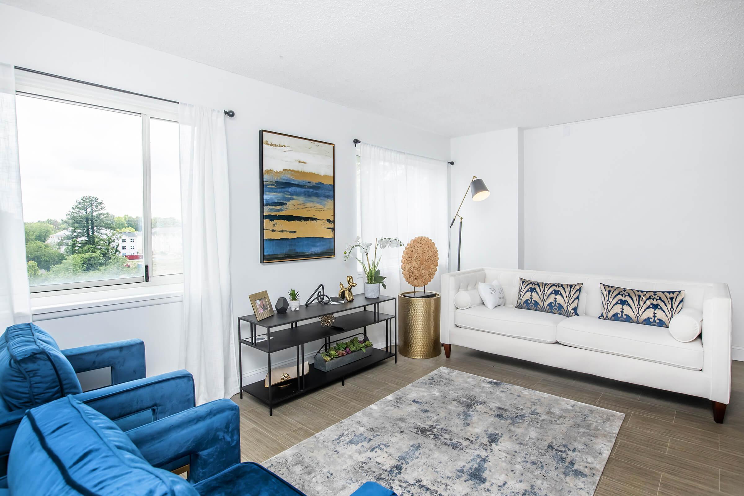 A bright, modern living room featuring a white sofa with blue patterned cushions, two blue armchairs, a decorative gold globe, and a stylish black shelving unit displaying plants and decor. Large windows let in natural light, and a light blue area rug complements the space.
