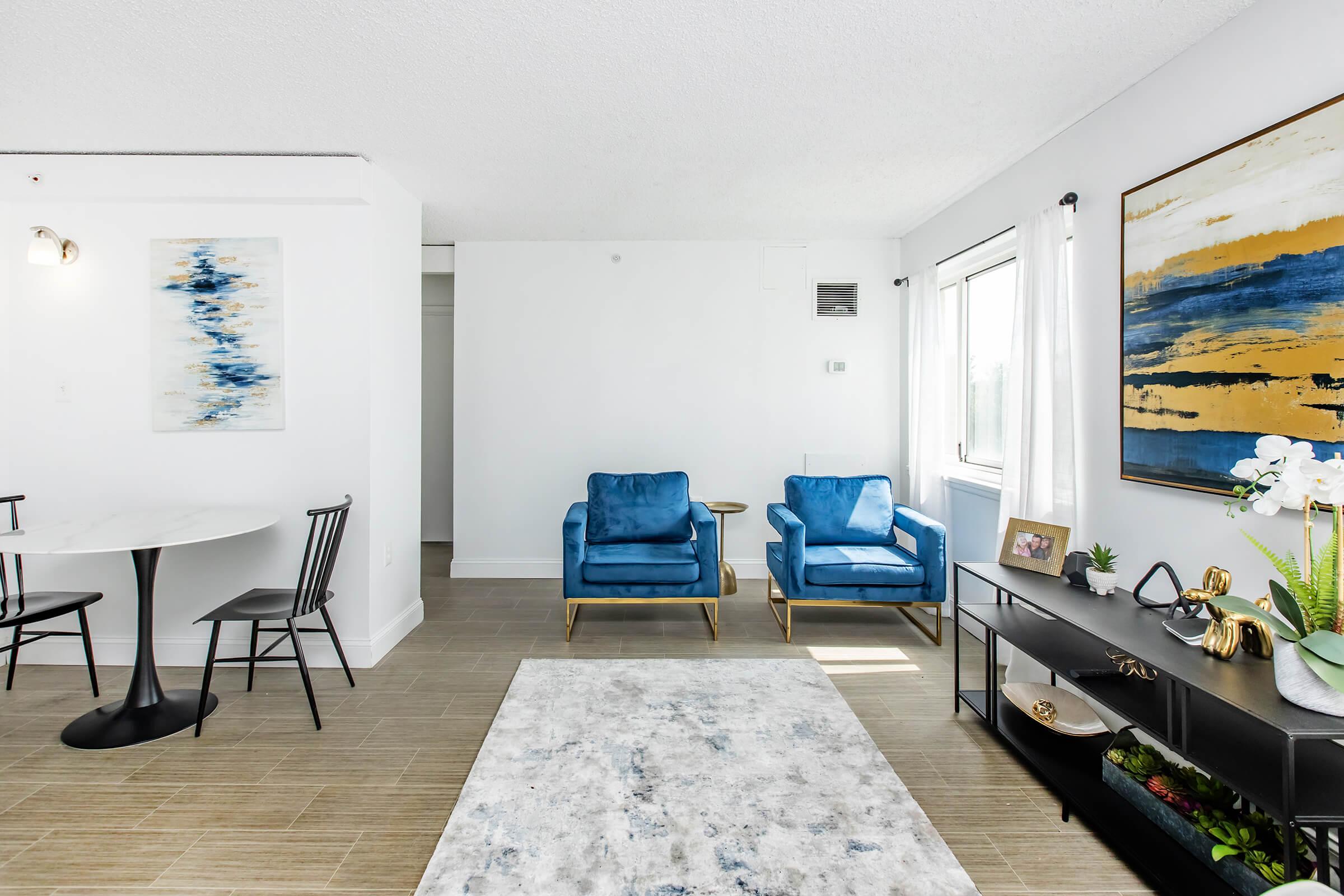 A modern living room featuring two blue armchairs, a round dining table with black chairs, and a decorative black console table. The space has light-colored walls and a neutral rug, with a large window allowing natural light. A piece of abstract art and a potted orchid add a touch of elegance.