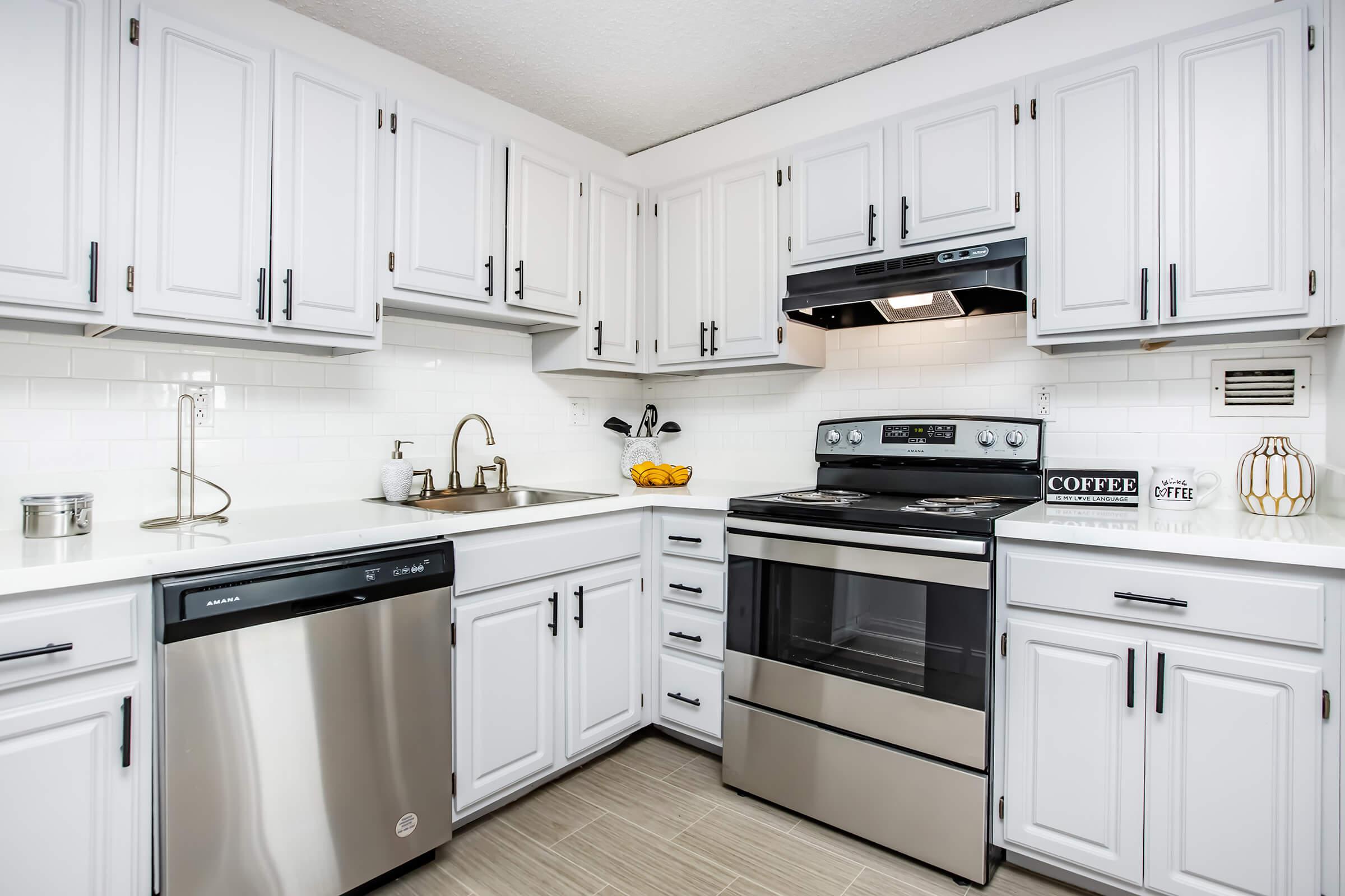 A modern kitchen featuring white cabinetry, stainless steel appliances, a black stove with an overhead exhaust fan, and a double sink. The countertop is clean and organized, with a dishwasher and decorative items, including a coffee jar and a bowl of fruit. The flooring is light-colored tiles, creating a bright and airy atmosphere.