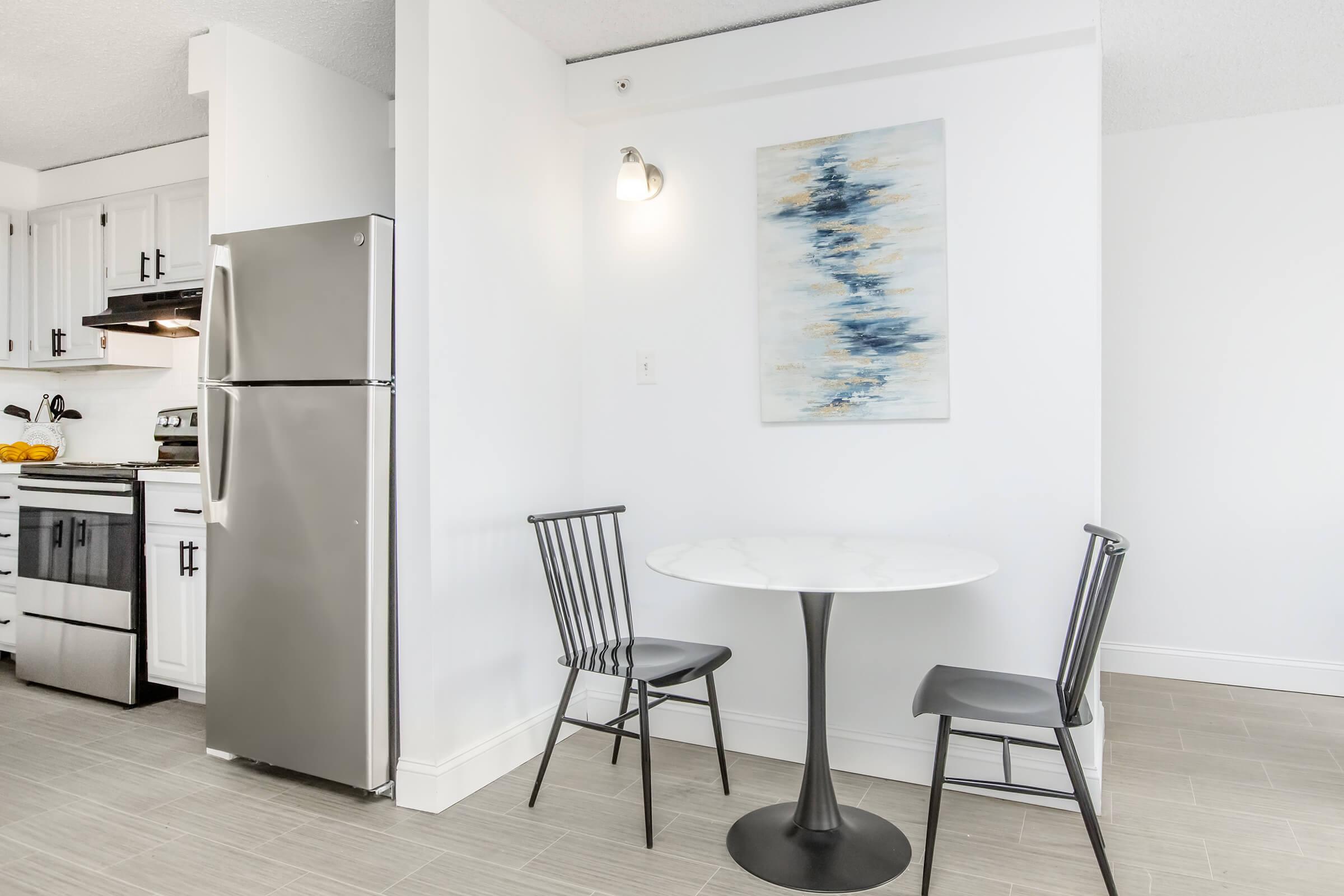 A small dining area featuring a round white marble table with two black chairs. In the background, there is a silver refrigerator and a small kitchen area with white cabinets. A wall painting with abstract blue and gold strokes adds a decorative touch to the space. The overall atmosphere is modern and bright.