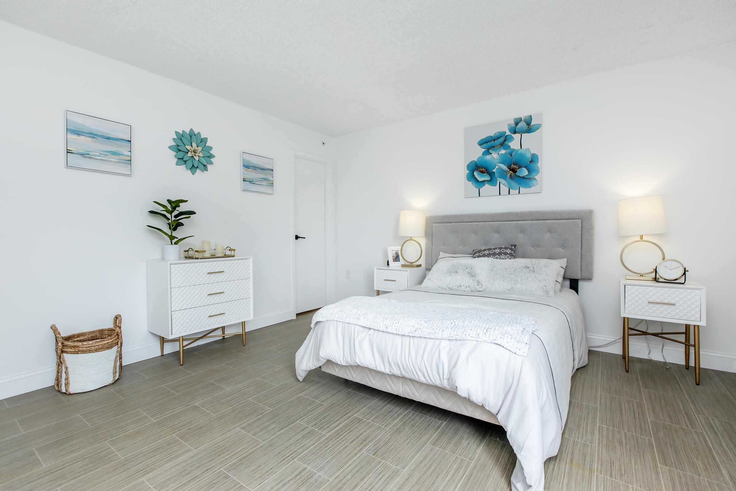 A bright, modern bedroom featuring a gray upholstered bed with white bedding. The room includes two small bedside tables with decorative lamps, a piece of artwork with blue flowers on the wall, and two framed coastal images. A plant adds a touch of greenery, and light-colored flooring completes the serene atmosphere.