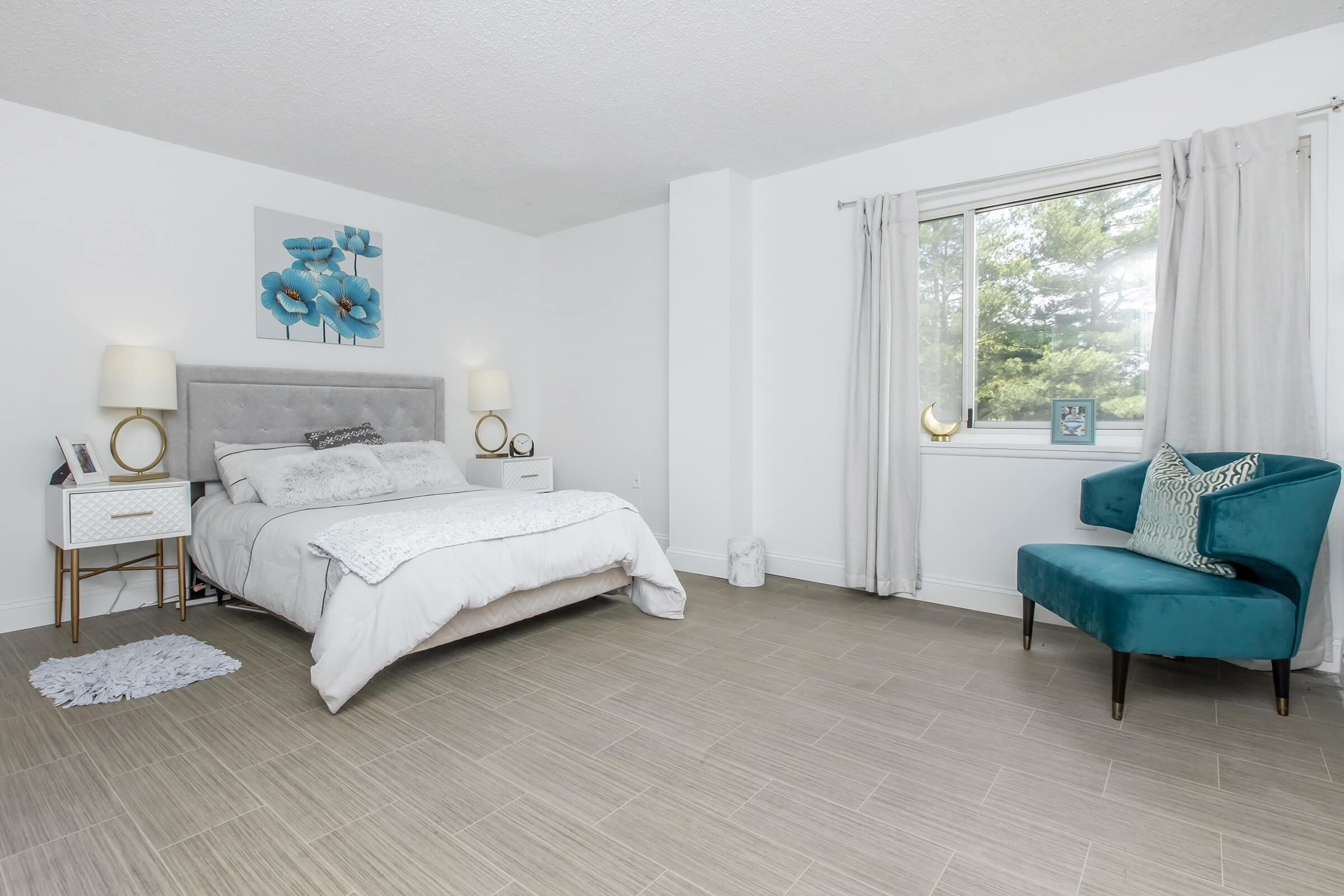 A modern bedroom featuring a comfortable bed with gray and white bedding, two bedside lamps, a decorative flower painting on the wall, a window with sheer curtains, and a cozy blue armchair positioned nearby. The floor is tiled, and the decor is simple and elegant.