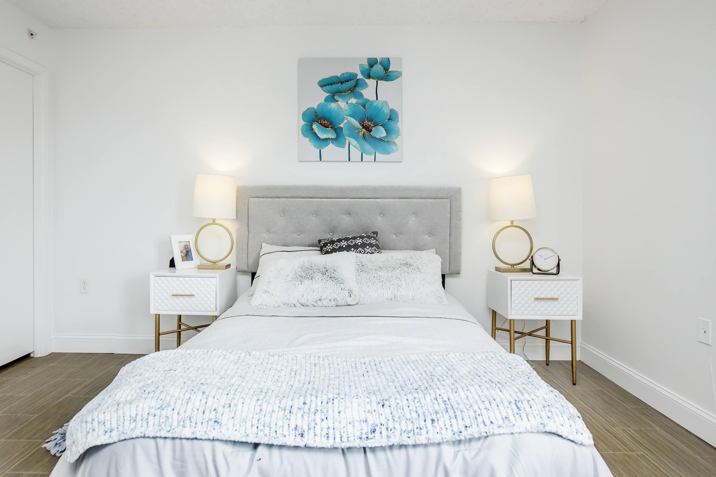 A modern bedroom featuring a light gray upholstered bed with a decorative white and blue throw blanket. Flanking the bed are two white nightstands with gold legs, each with a lamp and a clock. A piece of artwork with blue flowers is mounted above the bed, against a white wall. The flooring is light brown tiles.
