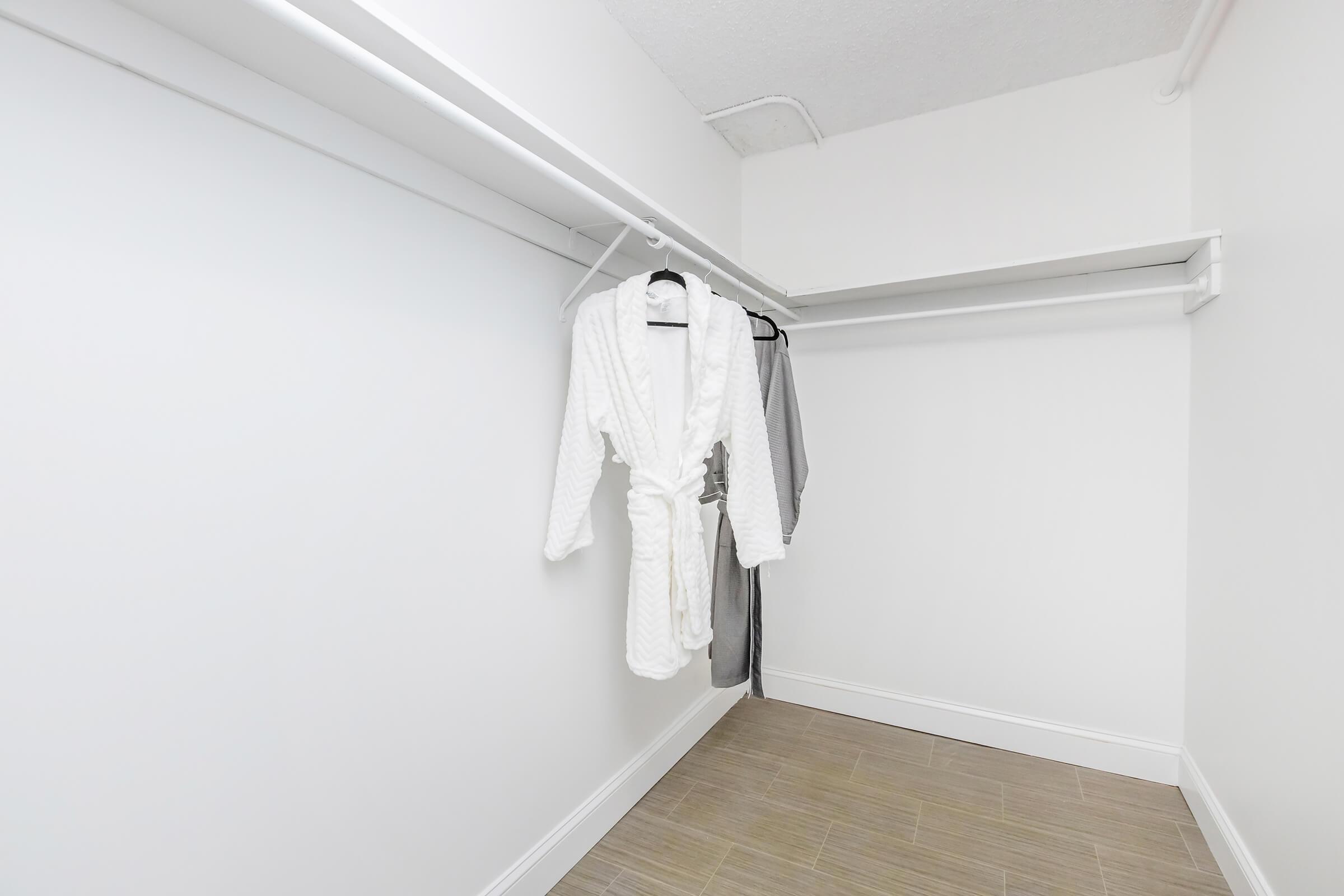 A minimalist closet featuring a white bathrobe hanging on a hanger, with gray clothing also displayed on a separate hanger. The walls are white, and the flooring is light-colored tile. The space is bright and uncluttered.