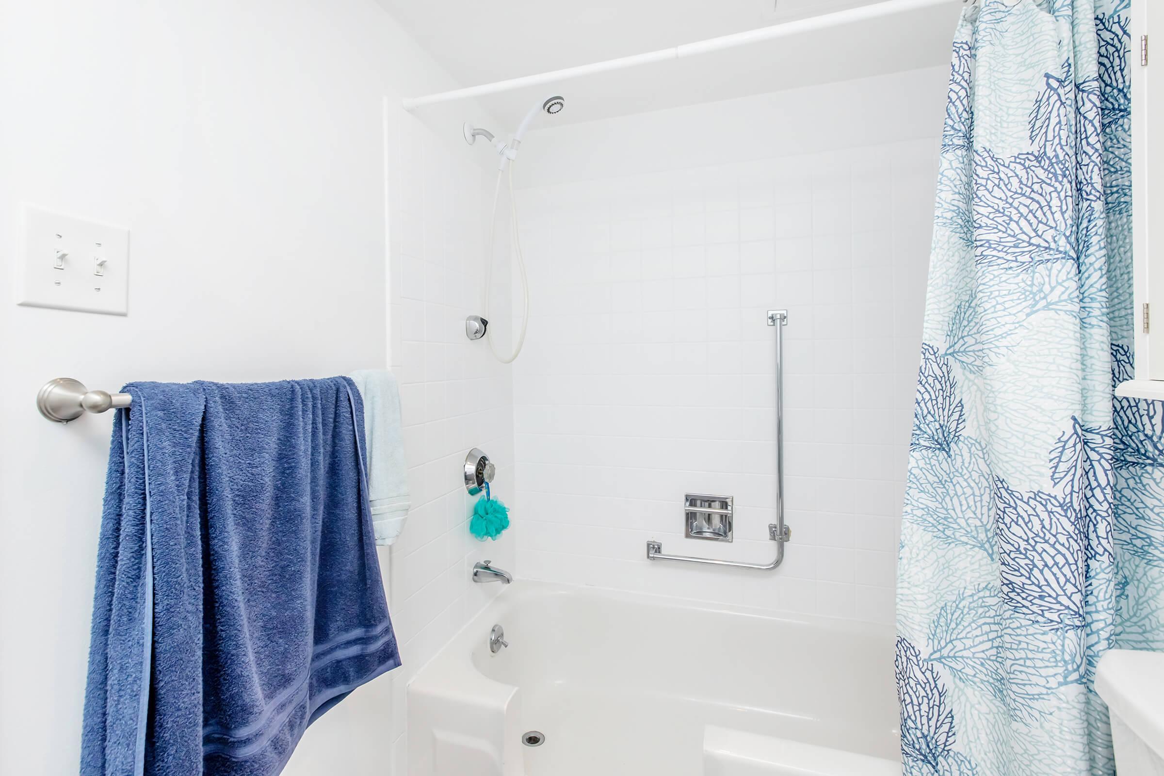 A clean and bright bathroom featuring a white tub with a shower, a blue coral-patterned shower curtain, and a blue towel hanging on a rack. The walls are white, creating a fresh atmosphere, and a showerhead is installed above the tub.