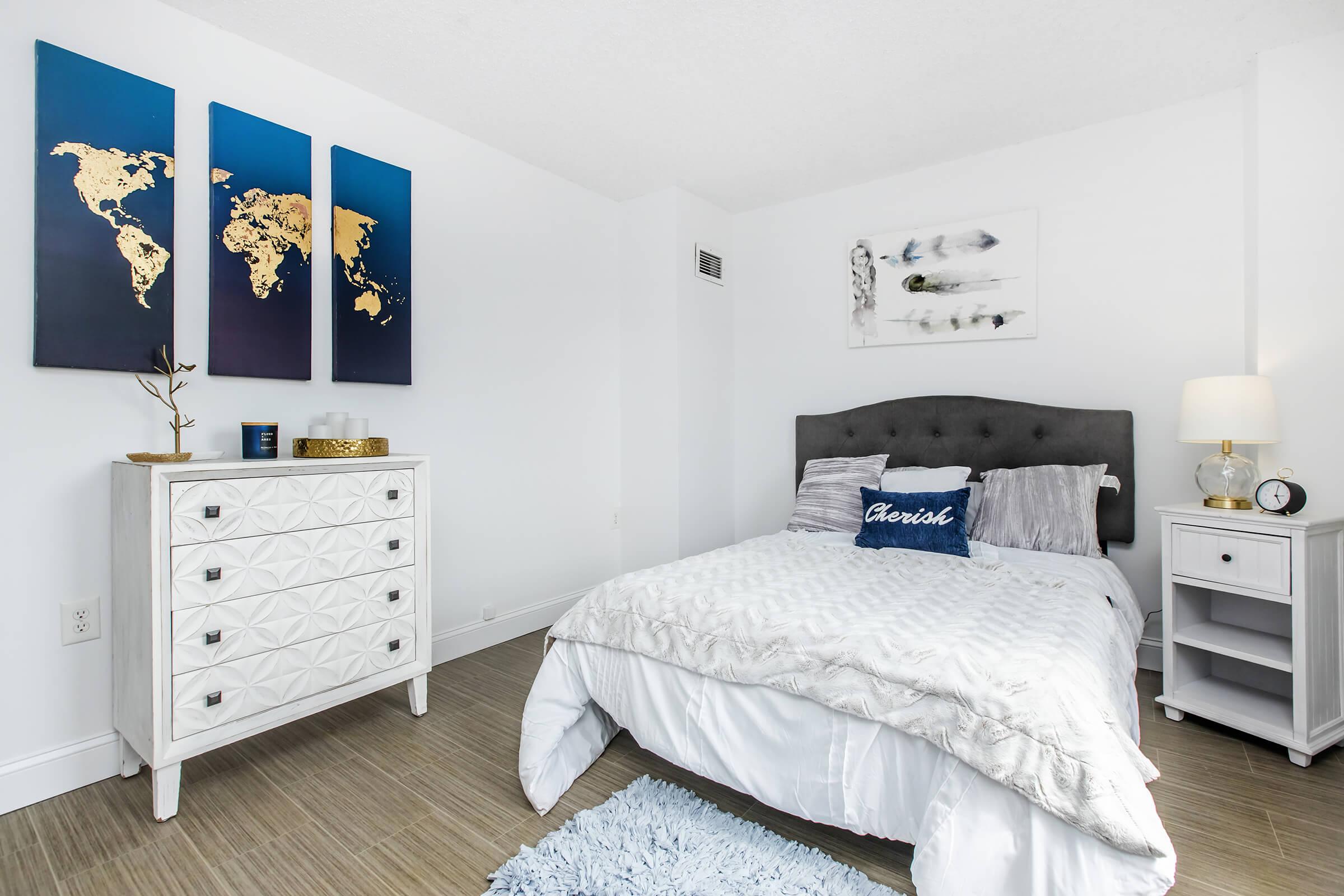A cozy bedroom featuring a bed with gray and white bedding, a decorative pillow that says "Aesthetic," and a stylish gray upholstered headboard. The room includes a modern white dresser with geometric patterns, a bedside table with a lamp and clock, and three gold-tone world map art pieces on the wall.