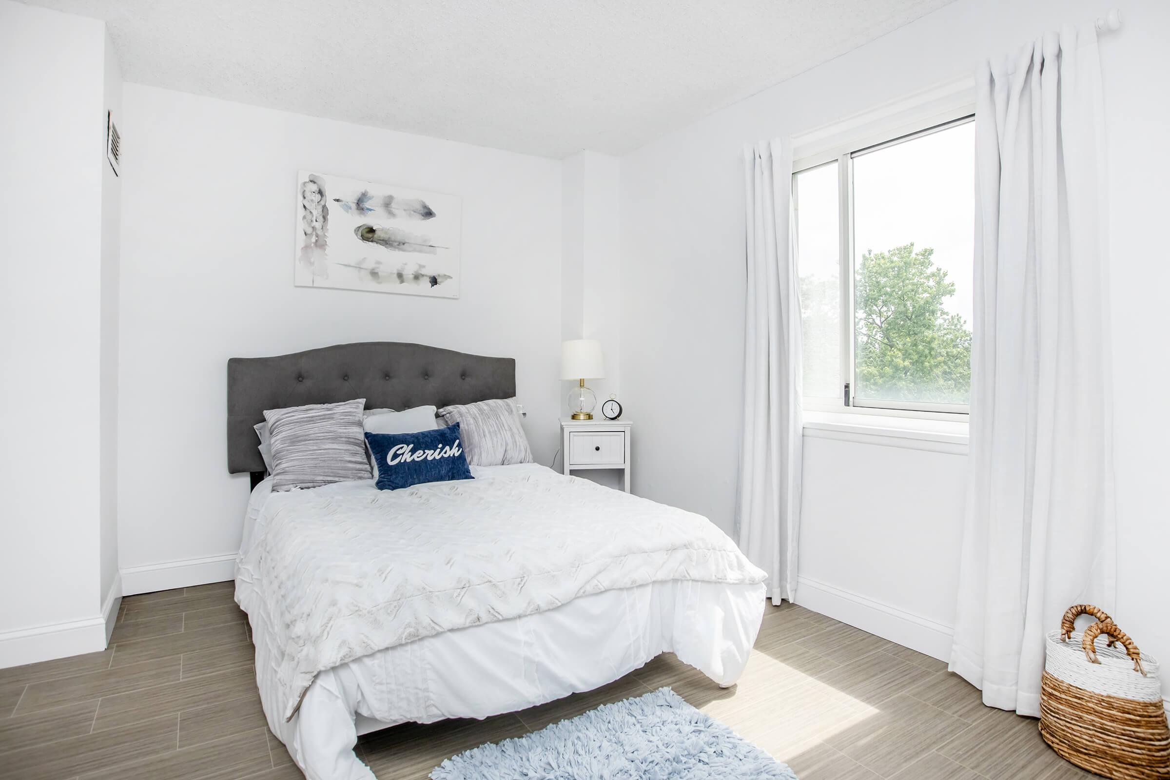 A cozy bedroom featuring a queen-sized bed with a light gray upholstered headboard and decorative pillows, a small nightstand with a lamp, and a wall art piece. There is a window with white curtains allowing natural light, and a woven basket in the corner adds a touch of texture to the space.