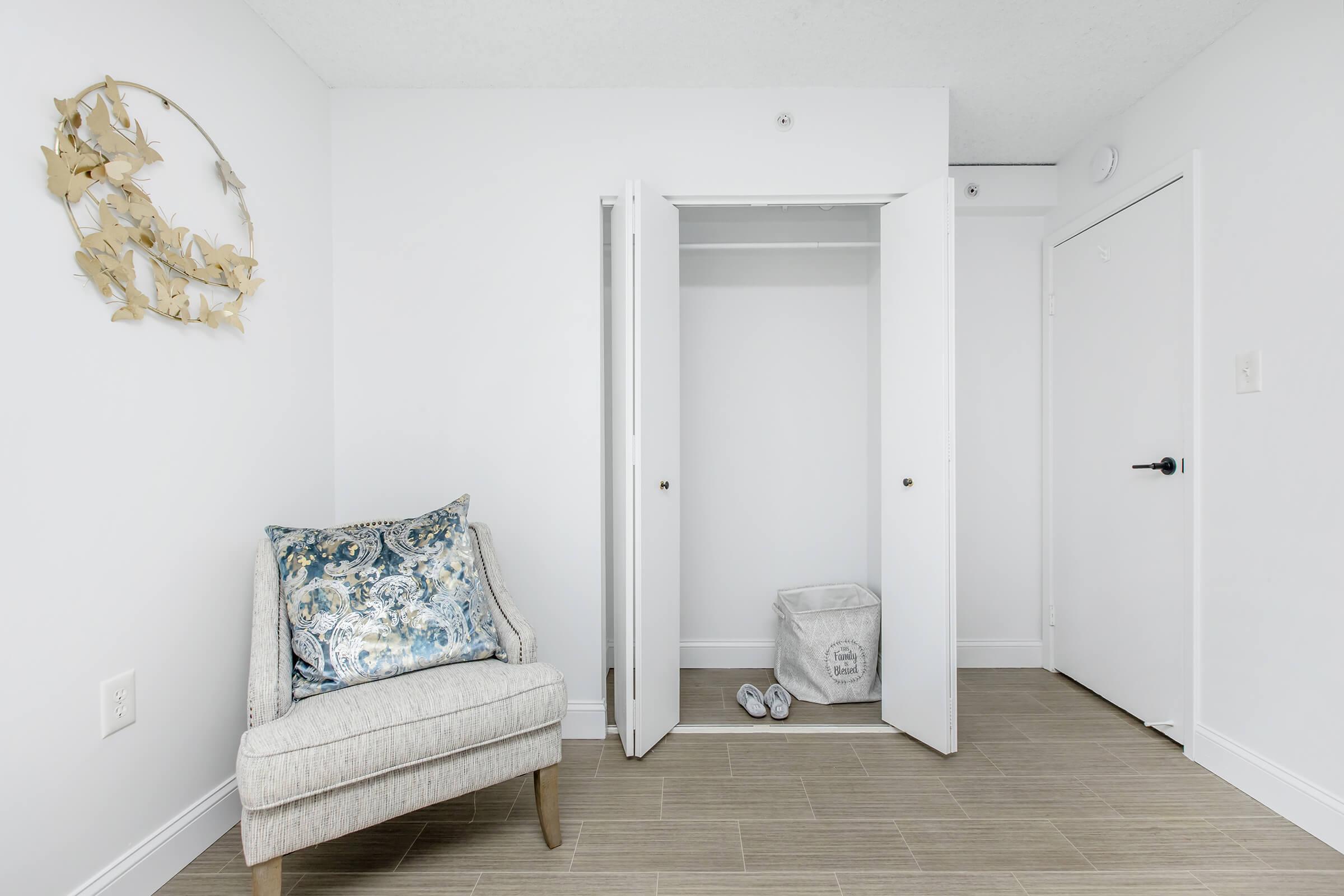 A bright, minimalist room with light-colored walls and floor. A comfortable chair with a decorative pillow is positioned next to an open closet featuring white doors. Inside the closet is a laundry basket, and there's a closed door on the right. The space is uncluttered and modern, creating a serene atmosphere.