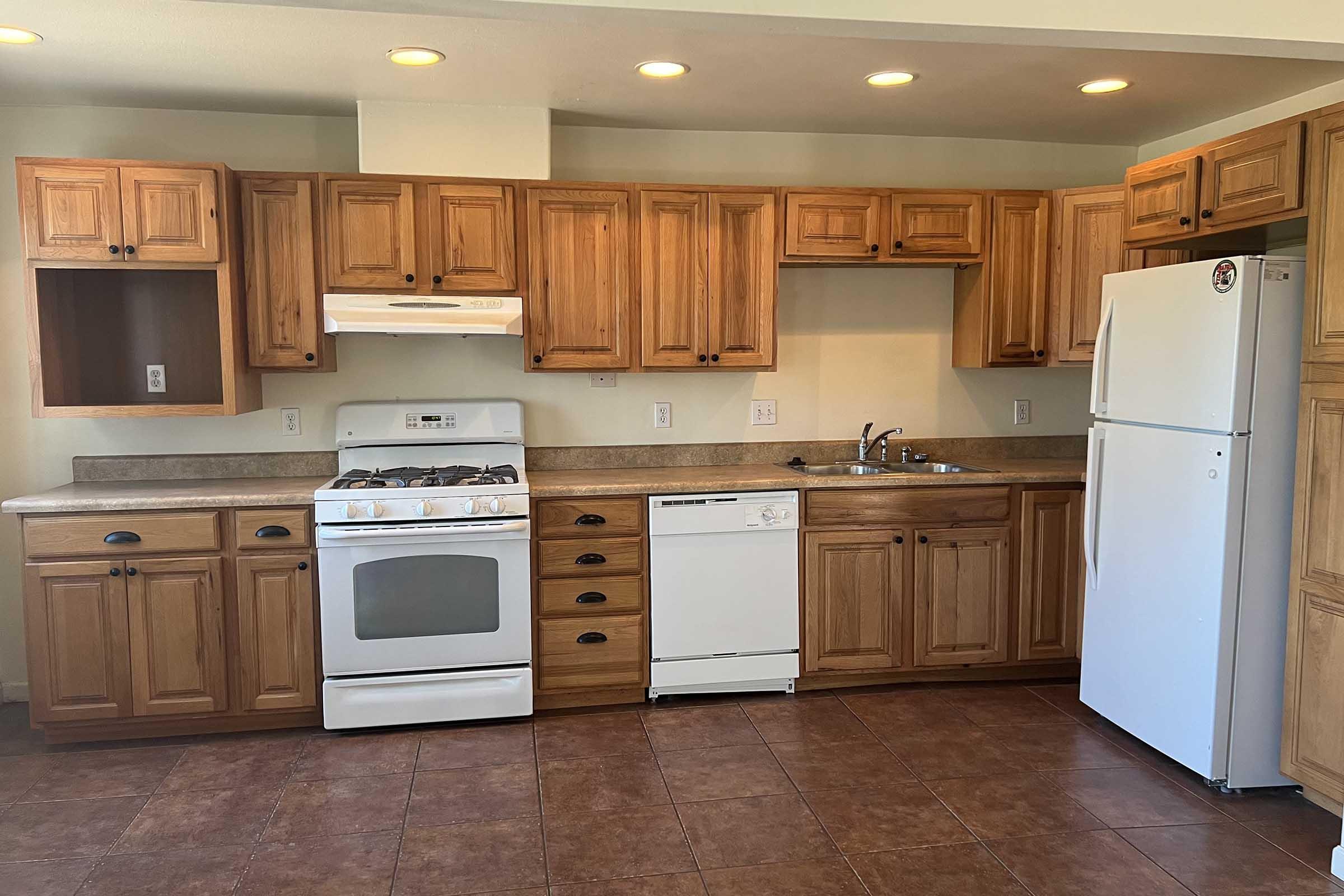 A kitchen with wooden cabinets, a gas stove, a dishwasher, and a refrigerator. The countertop is a light color, and the floor consists of dark tiles. There is overhead lighting, and the walls are painted a neutral tone.