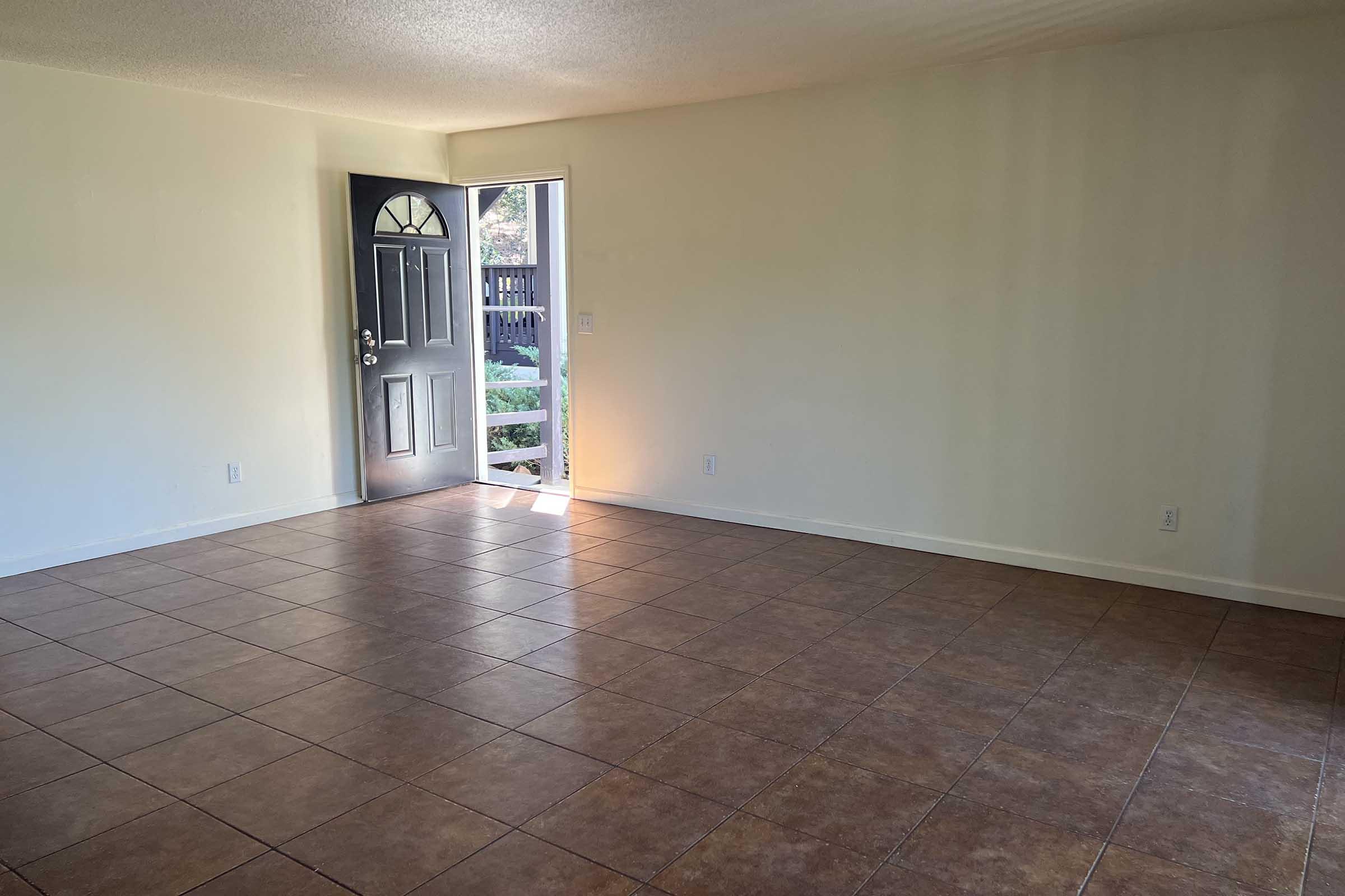 A spacious, unfurnished living room with tiled flooring and a neutral wall color. A door on the left leads outside, allowing natural light to enter. The room is empty, creating an open and airy feel, with a visible view of outdoor greenery through the door.