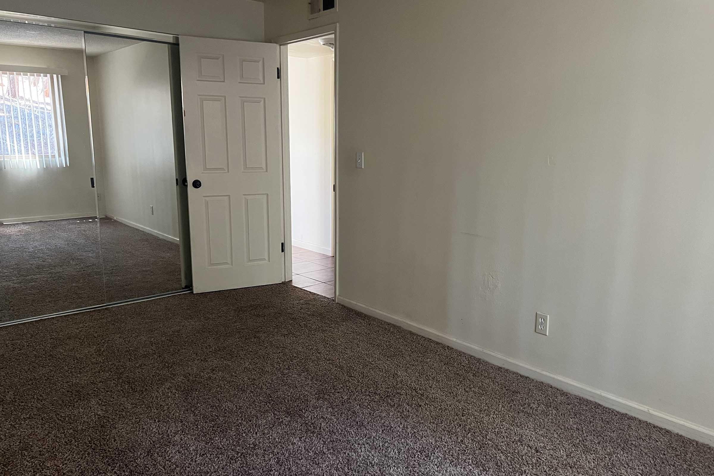 A spacious room with light beige walls and a plush carpet. To the left, there is a large mirror sliding closet door, reflecting the room. A doorway leads to another space, visible in the background. Natural light filters in through a window, creating a bright and open atmosphere.