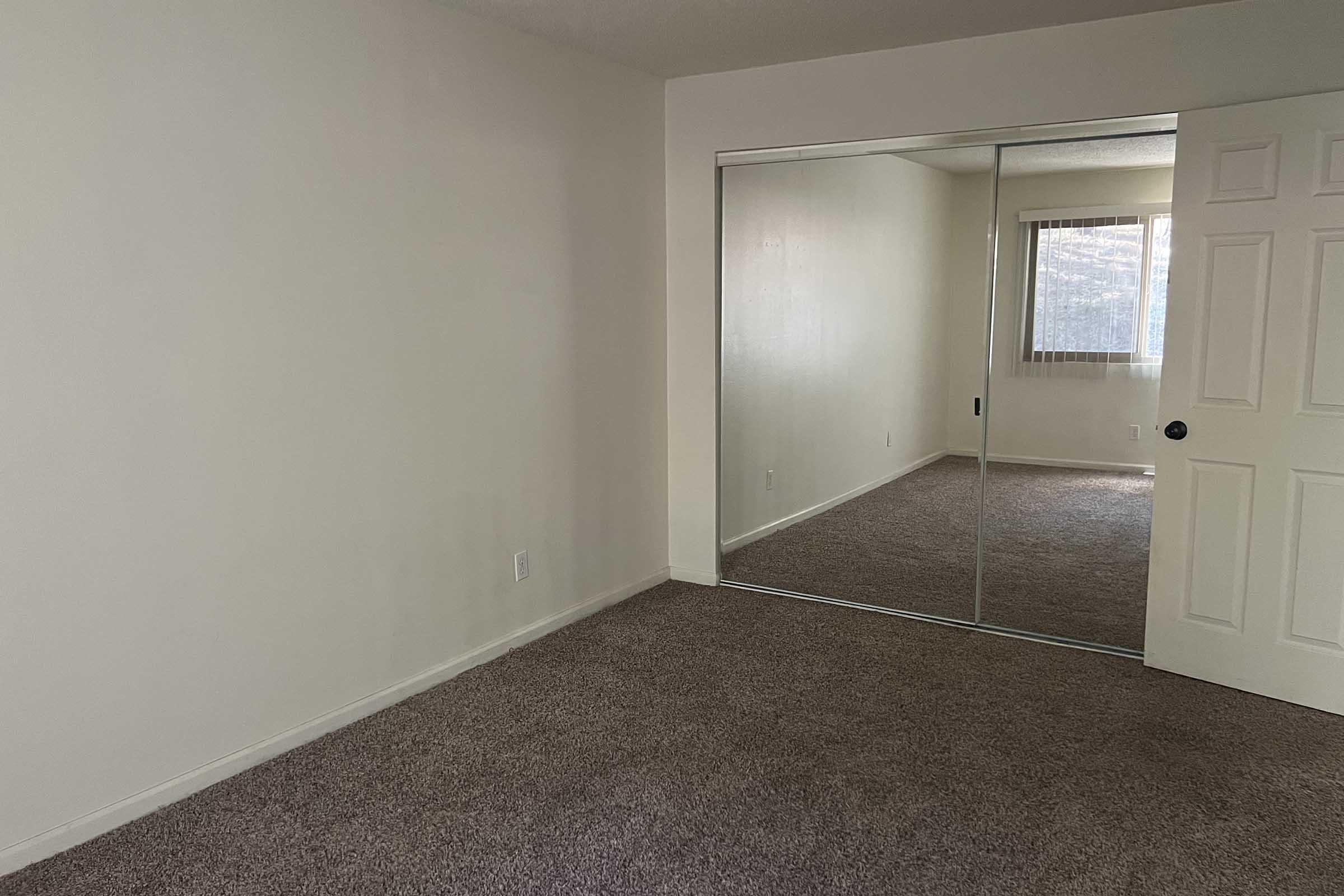 A light-colored room with beige carpet and white walls. There is a large mirrored closet on one side. A window with a sheer curtain allows natural light to enter, and the opposite wall has a closed door. The space appears empty and well-lit.