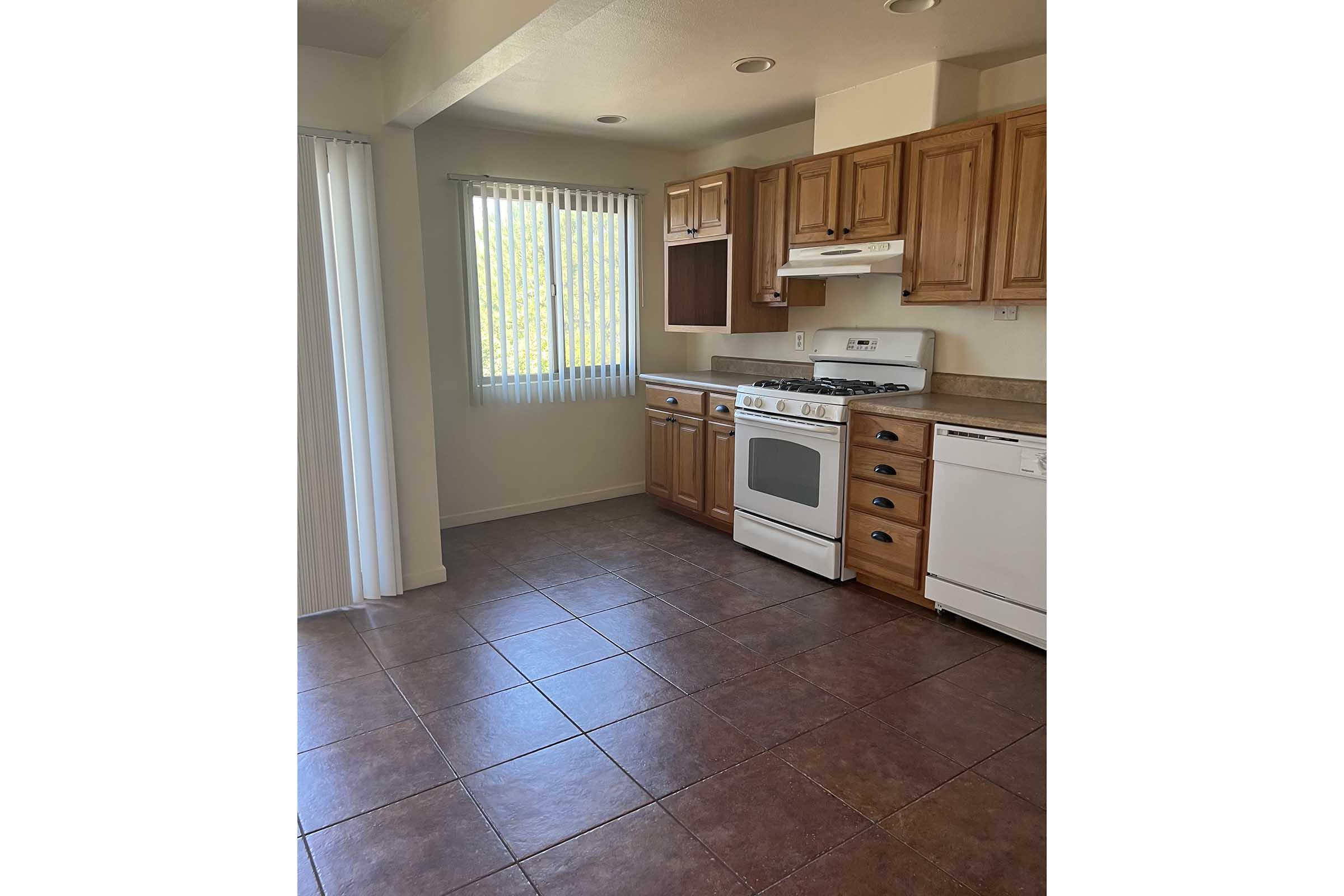 Bright kitchen featuring wooden cabinets, a white stove and oven, and a dishwasher. Light streaming in through a window with vertical blinds. The floor is tiled with large brown tiles, and the overall space is open and inviting, showcasing an organized and clean layout.