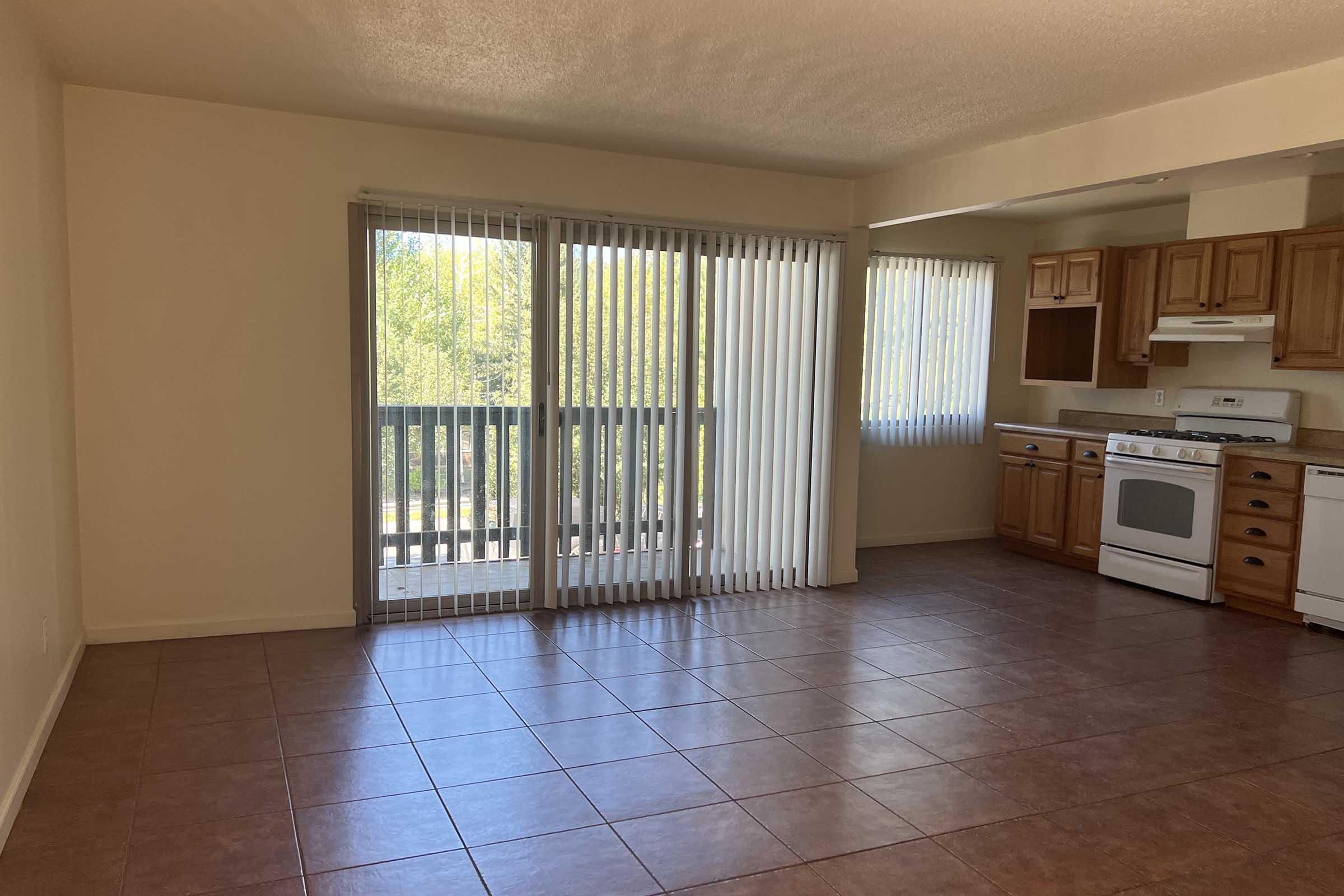 A bright, spacious living area with large sliding glass doors leading to a balcony. The room features tiled flooring, vertical blinds, and an adjoining kitchen with wooden cabinets and a white stove. Natural light floods in from the windows, creating a warm atmosphere.