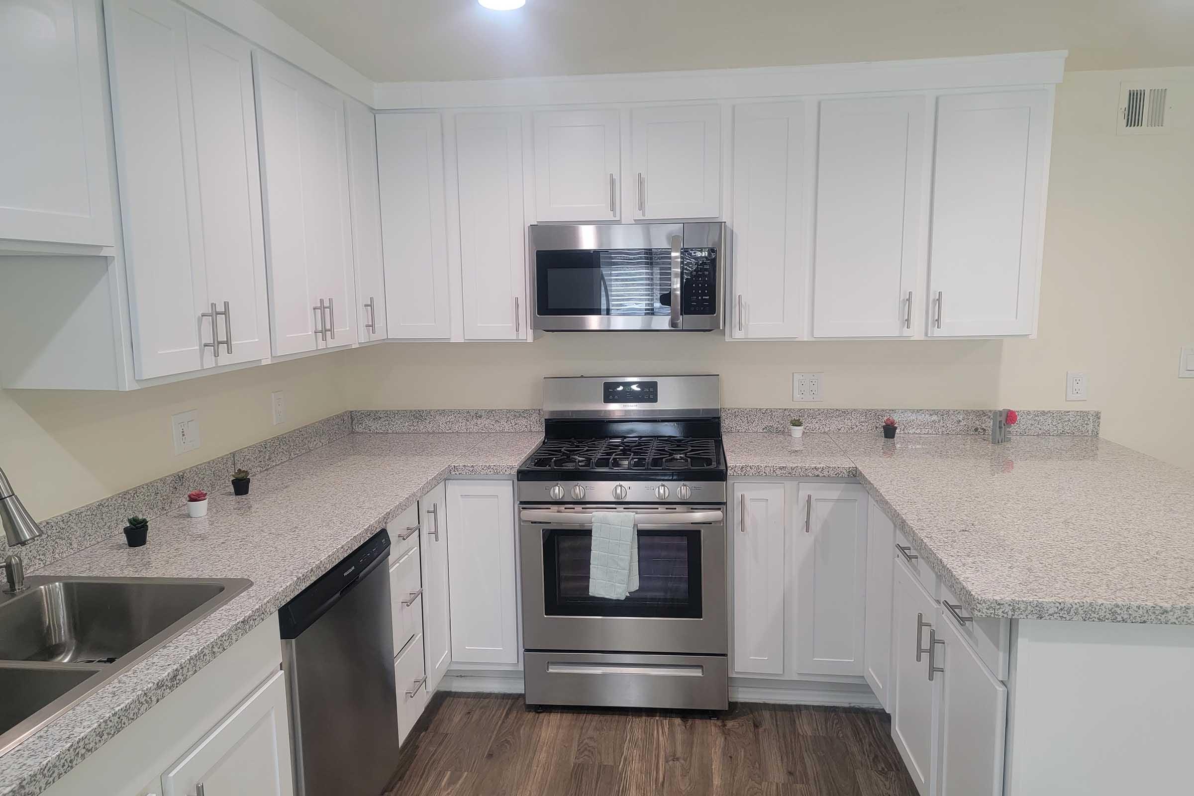 A modern kitchen featuring white cabinets, granite countertops, and stainless steel appliances. It includes a gas stove and microwave, along with a sink and dishwasher. The space is well-lit, with a neutral color palette and minimal decor.