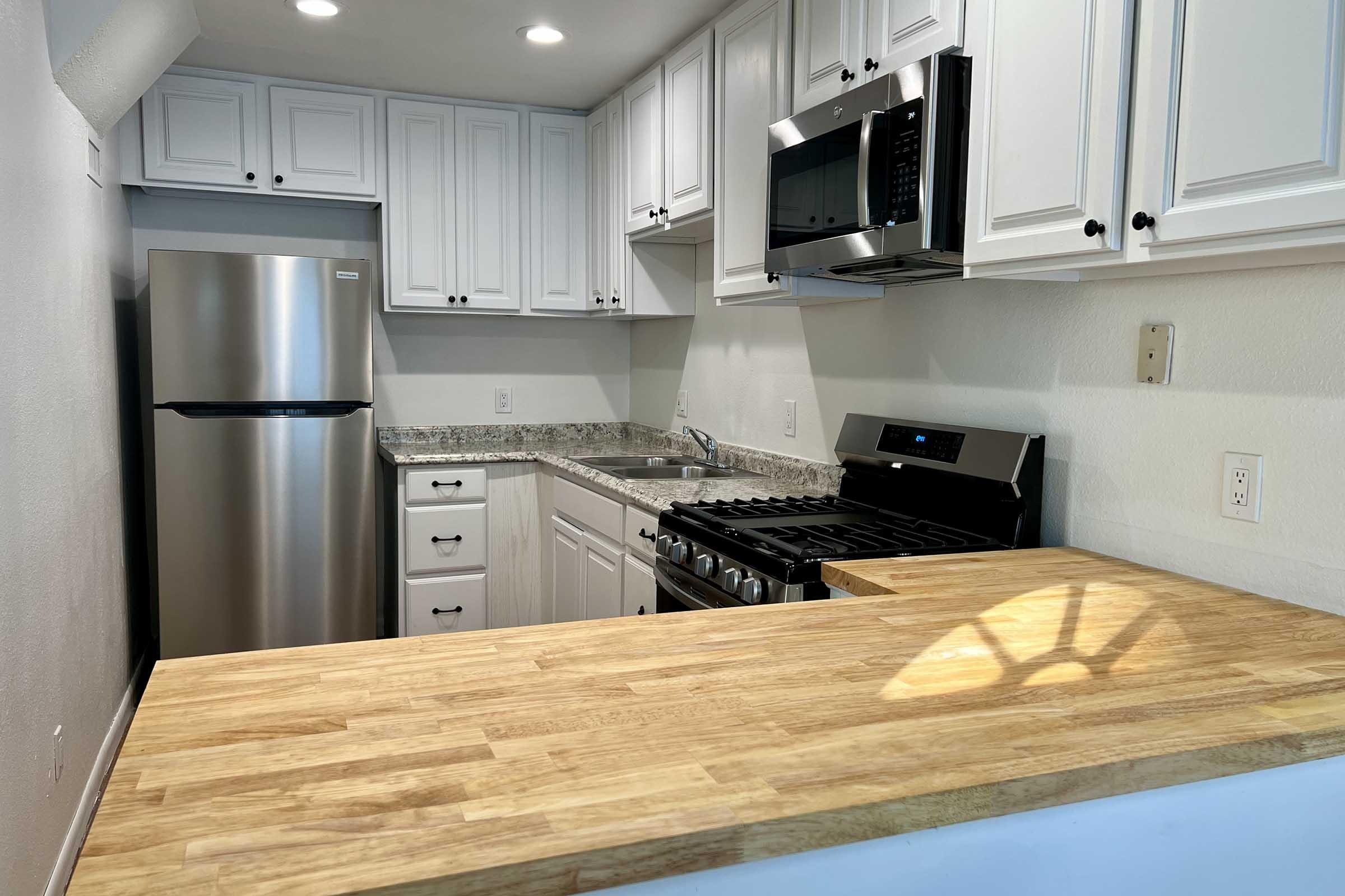 Modern kitchen featuring white cabinets, stainless steel appliances including a refrigerator and microwave, granite countertops, and a wooden breakfast bar. The kitchen is well-lit with recessed lighting and has a clean, minimalist design.