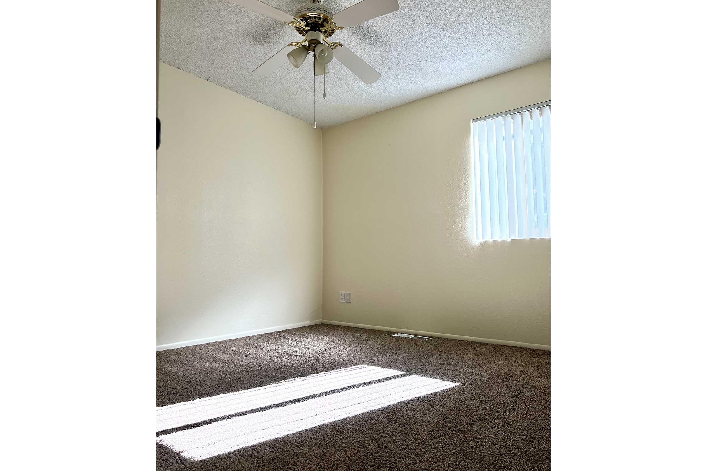 A small, empty room with light beige walls and a textured ceiling. A ceiling fan with three blades is mounted in the corner. A single window with vertical blinds allows natural light to stream in, casting shadows on the carpeted floor. The room appears bright and inviting, with no furniture present.