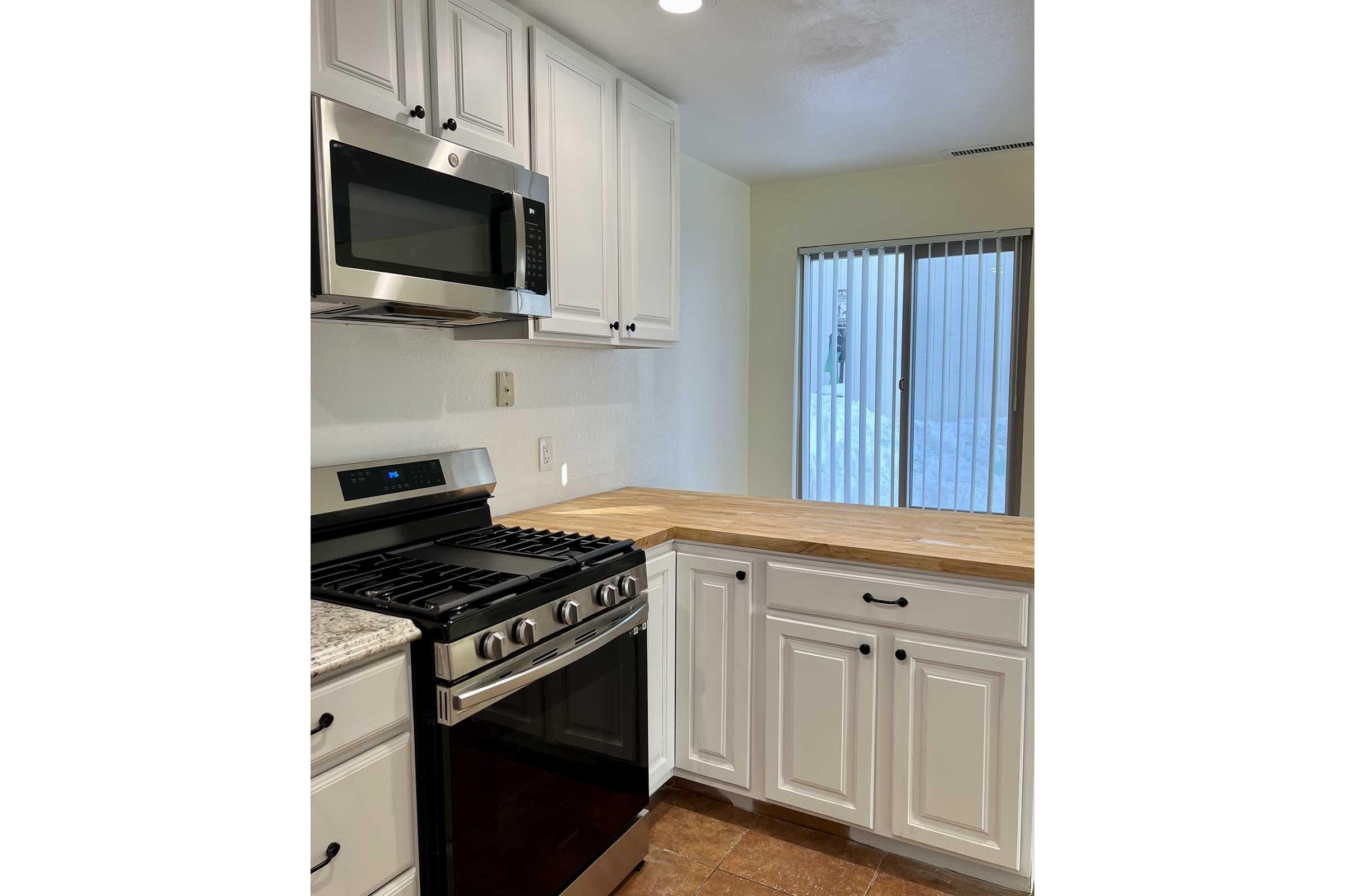 Modern kitchen featuring white cabinets, a stainless steel microwave, and a gas range with an oven. A wooden countertop extends from the cabinetry, and there's a window with vertical blinds allowing natural light. The flooring is tiled in a warm tone, creating a welcoming atmosphere.