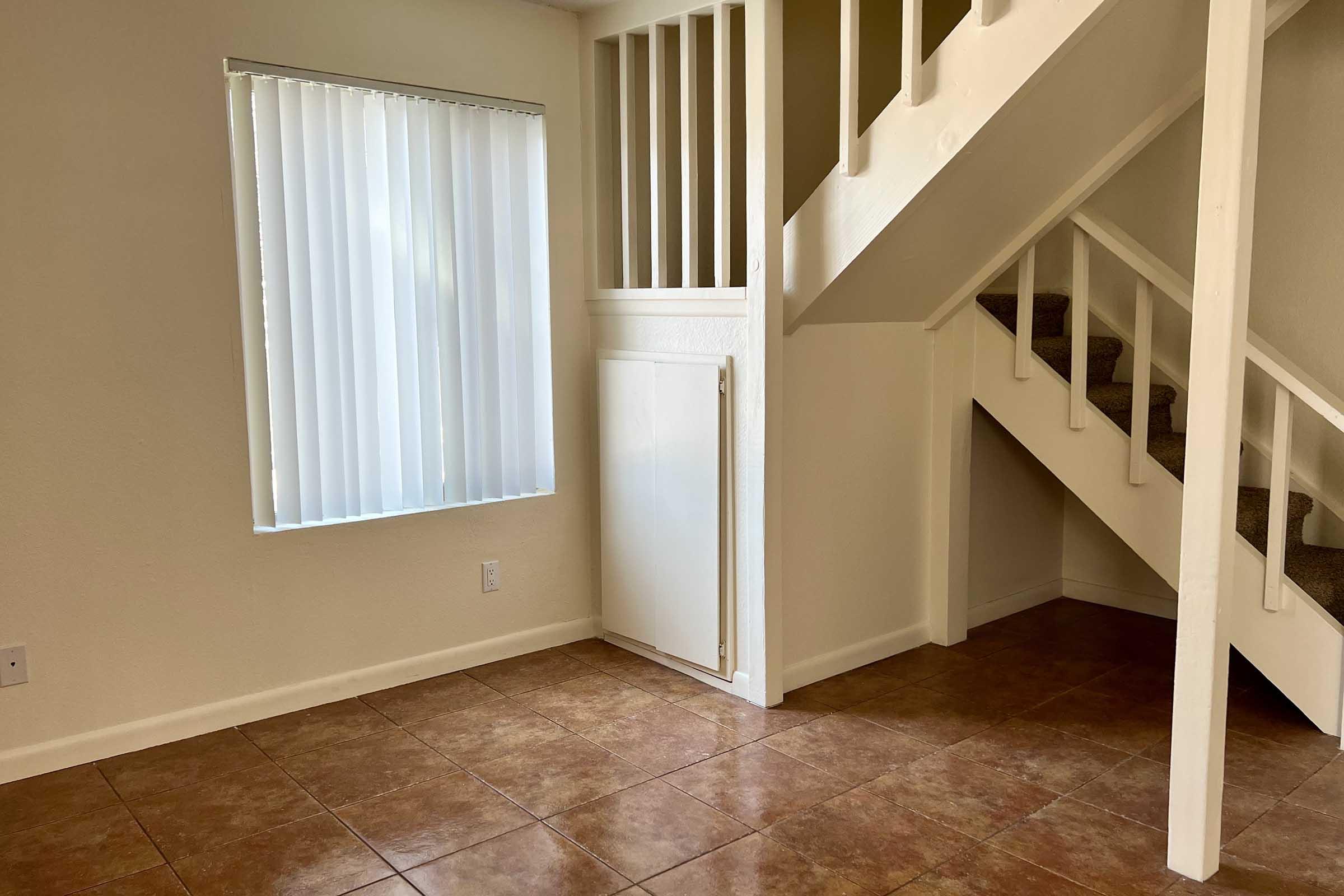 Interior view of a room featuring beige walls and tiled flooring. A staircase is visible in the corner, leading to an upper level. To the left, there is a window covered with vertical blinds, allowing natural light into the space. The room appears empty, providing a blank canvas for potential furnishing.