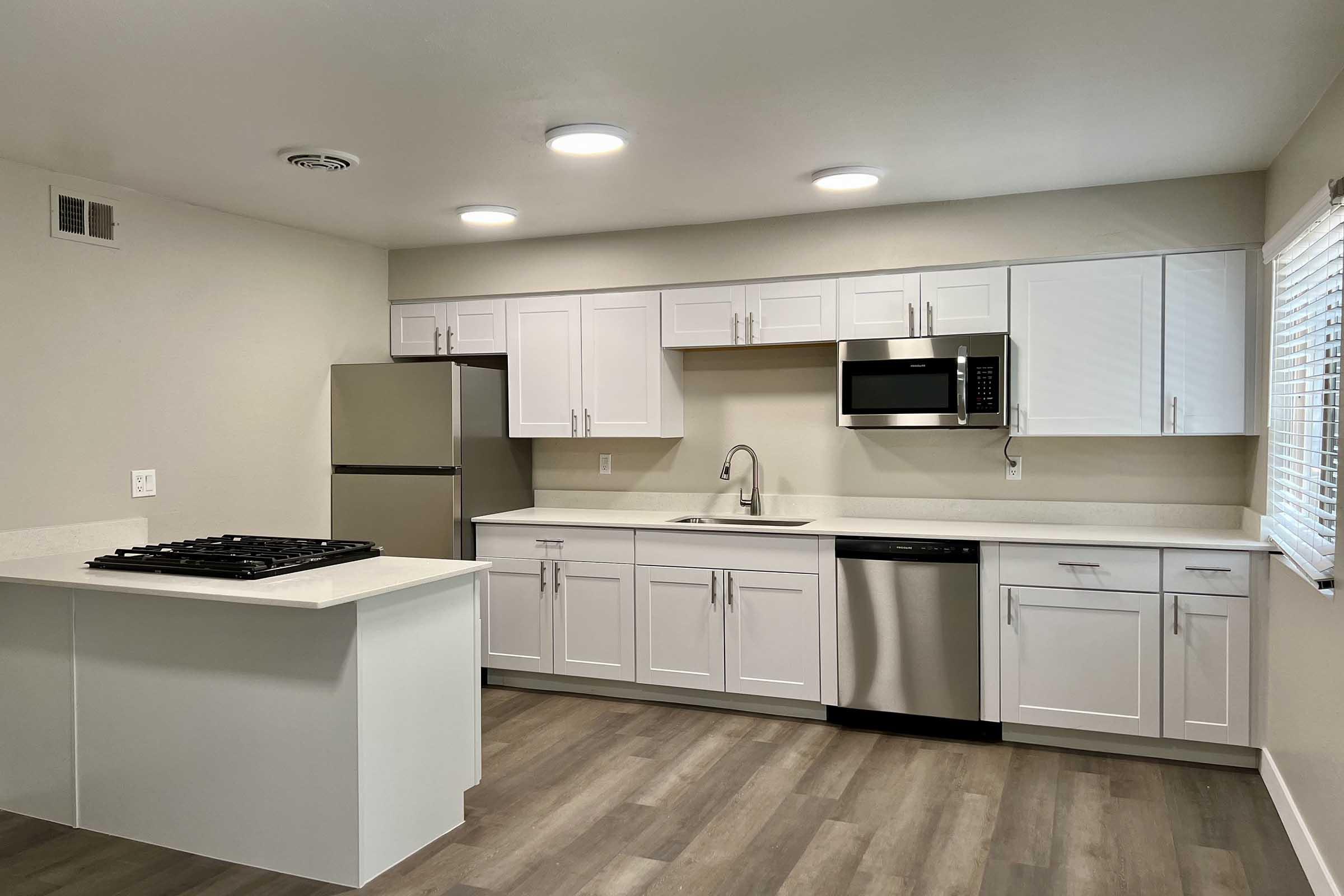 Modern kitchen featuring white cabinets, stainless steel appliances including a refrigerator, microwave, and dishwasher. A center island with a gas cooktop is present. The walls are a light color, and there is ample natural light coming from a window with blinds.