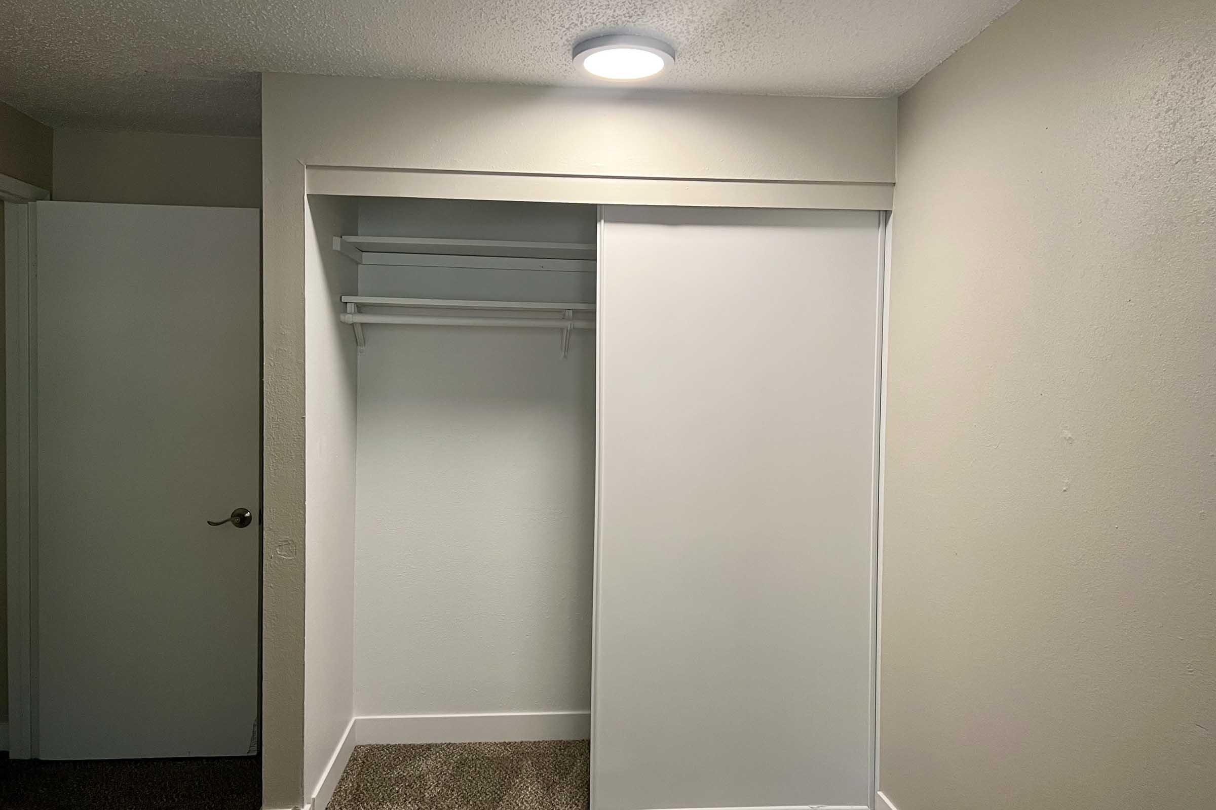 Interior view of a simple closet featuring a sliding white door, a shelf above, and an empty space below. The walls are painted light colors, and the floor is covered with carpet. A ceiling light is visible, providing illumination to the area.