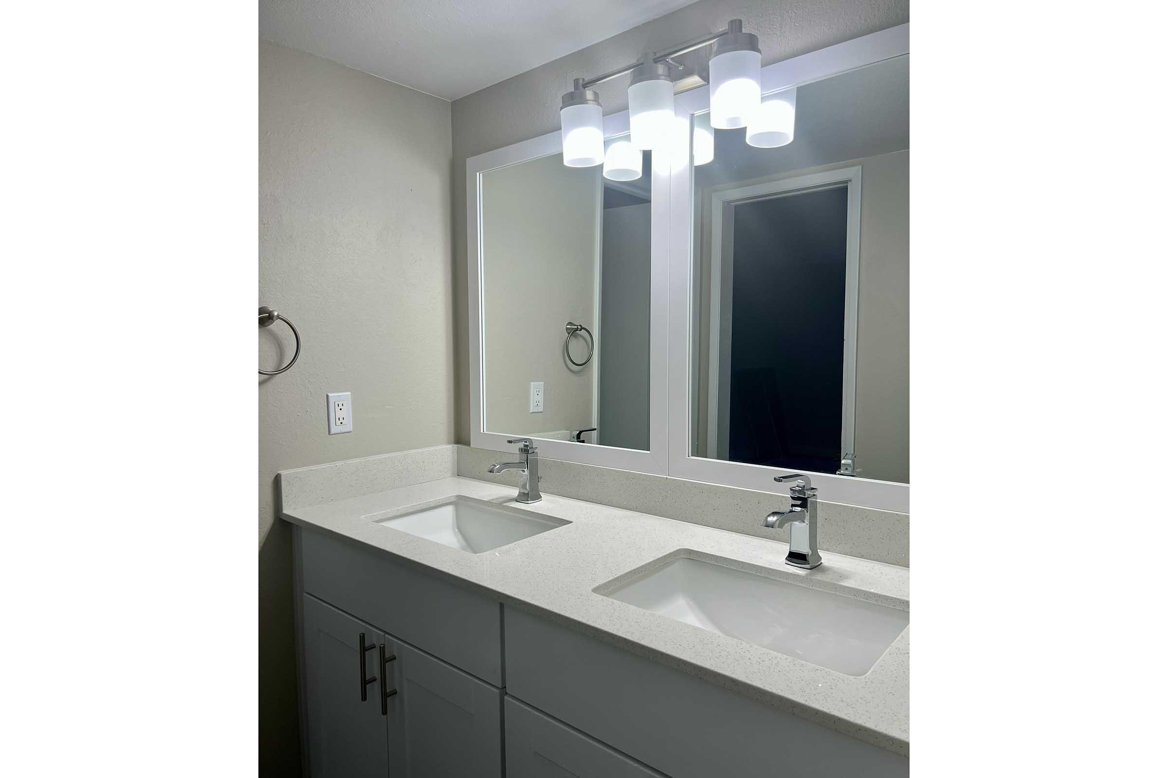 Modern bathroom with a double vanity featuring two rectangular sinks, sleek chrome faucets, and a light-colored countertop. Above the sinks, there are two large mirrors flanked by four round light fixtures. The walls are painted in a neutral color, creating a clean and inviting atmosphere.