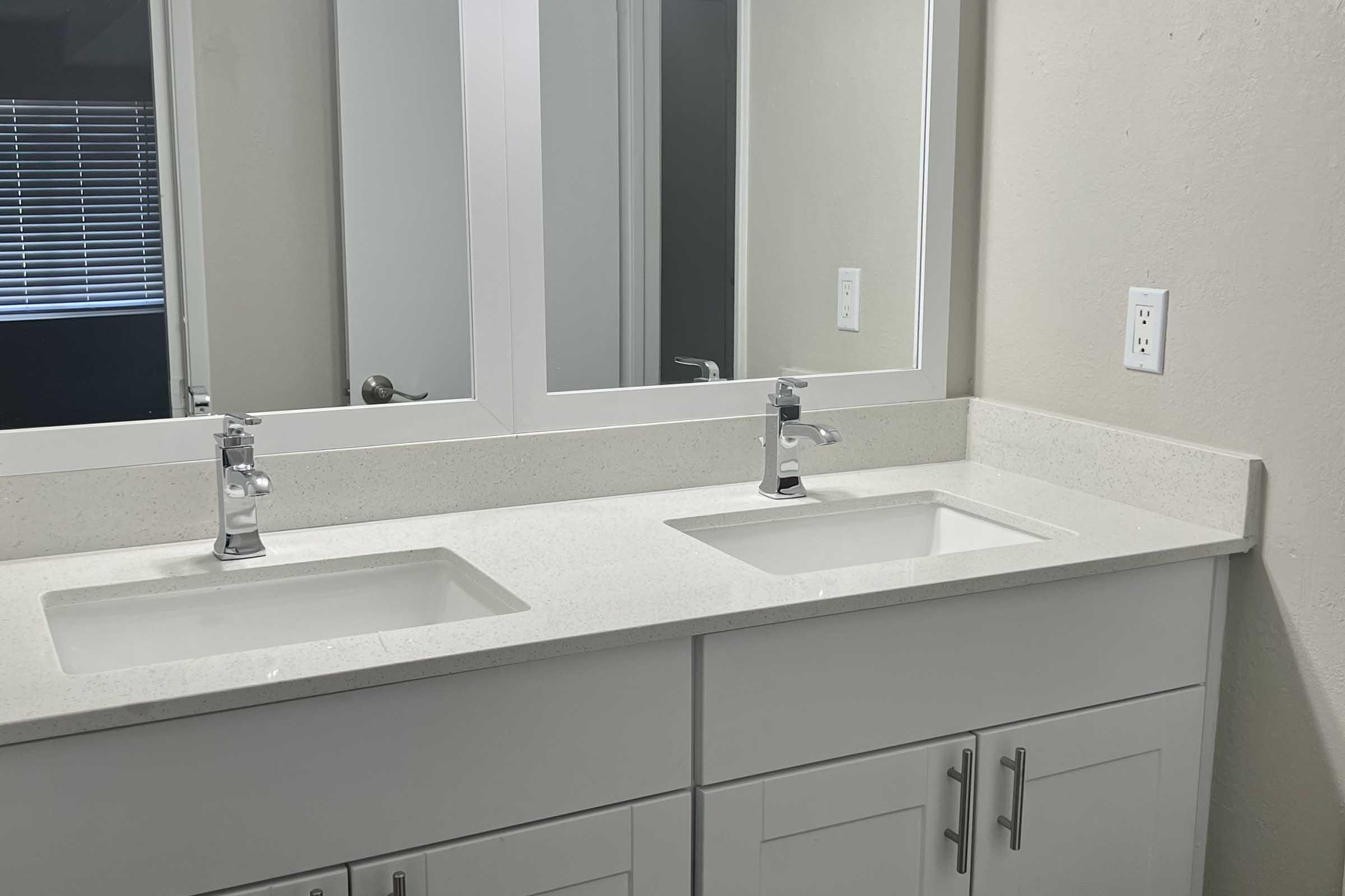 Modern bathroom vanity with a double sink and chrome faucets. The countertop is light-colored stone, and there are two mirrors above the sinks. The cabinetry below features a clean, minimalist design with white doors. Natural light is visible through a window in the background.