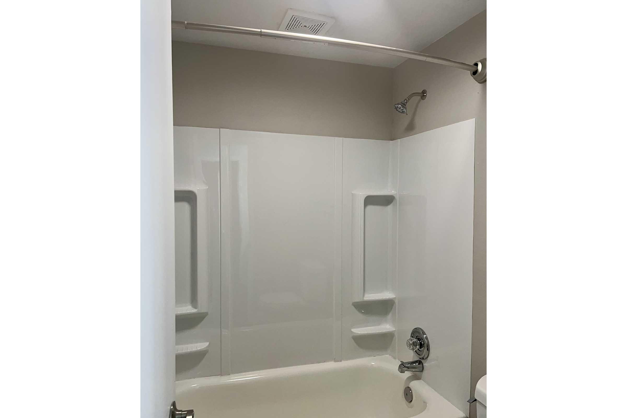 A clean, empty bathtub with a white acrylic surround. The showerhead is mounted on the wall, and there are built-in shelves for toiletries on both sides. The walls are painted a light color, contributing to a bright and spacious feel in the bathroom.